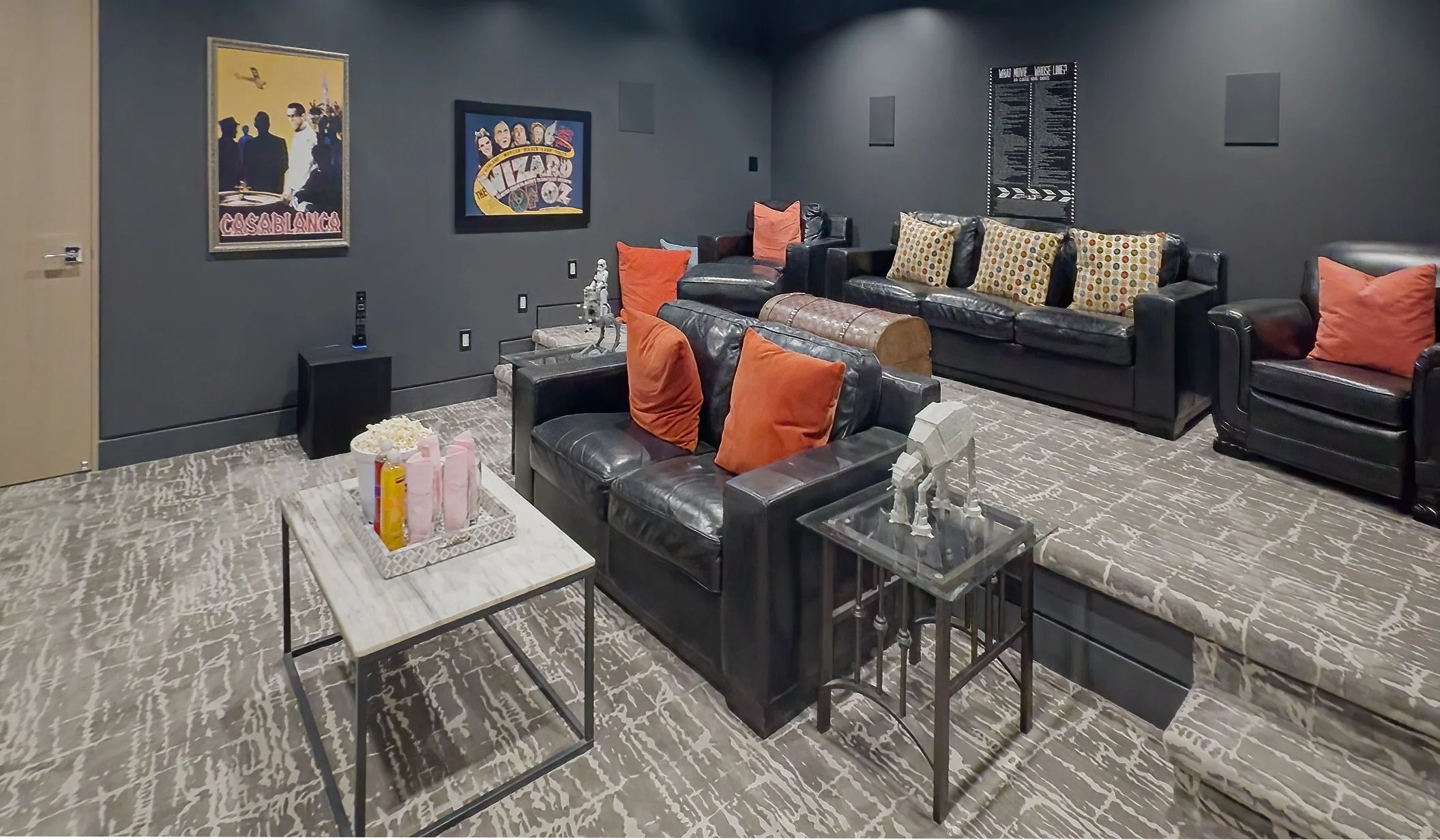 A cozy theater room with dark walls, black leather sofas with orange and patterned pillows, wall art posters, a small side table with decorations, a coffee table with snacks, and a plush carpet.