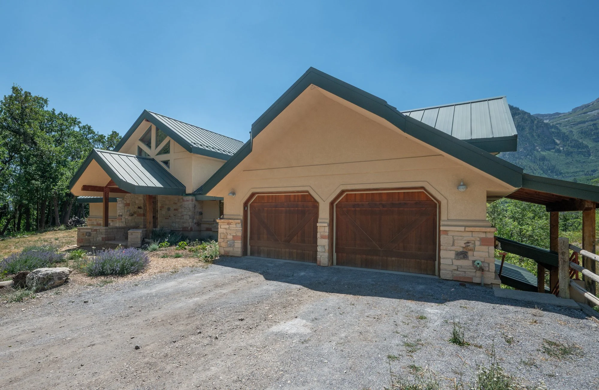 A house with a sloped green metal roof, stone accents, and a double wooden garage door, surrounded by trees and mountains under a clear blue sky.