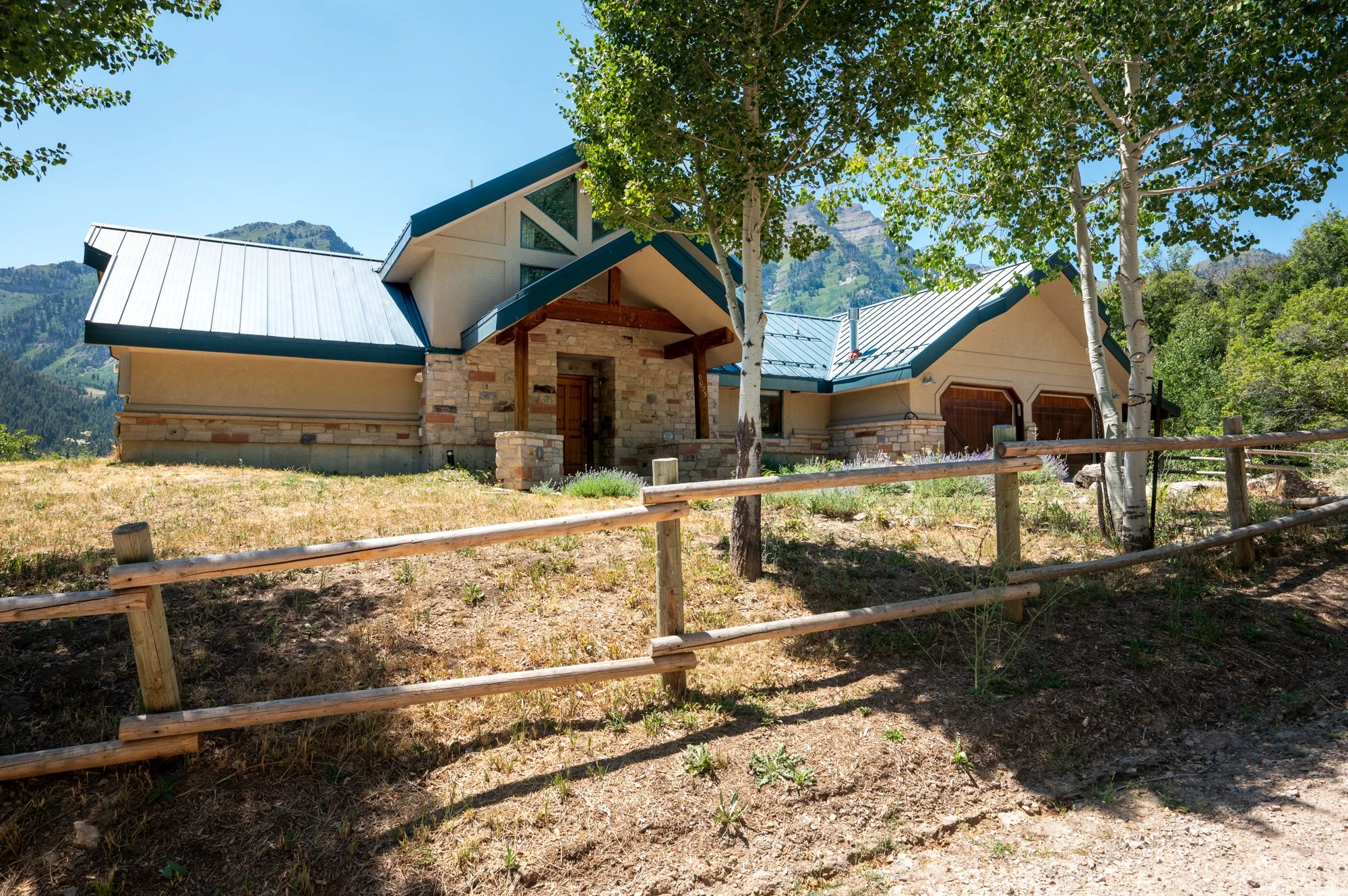 A house with a modern design, stone accents, and a blue metal roof, surrounded by trees and mountains, with a wooden fence in the foreground.