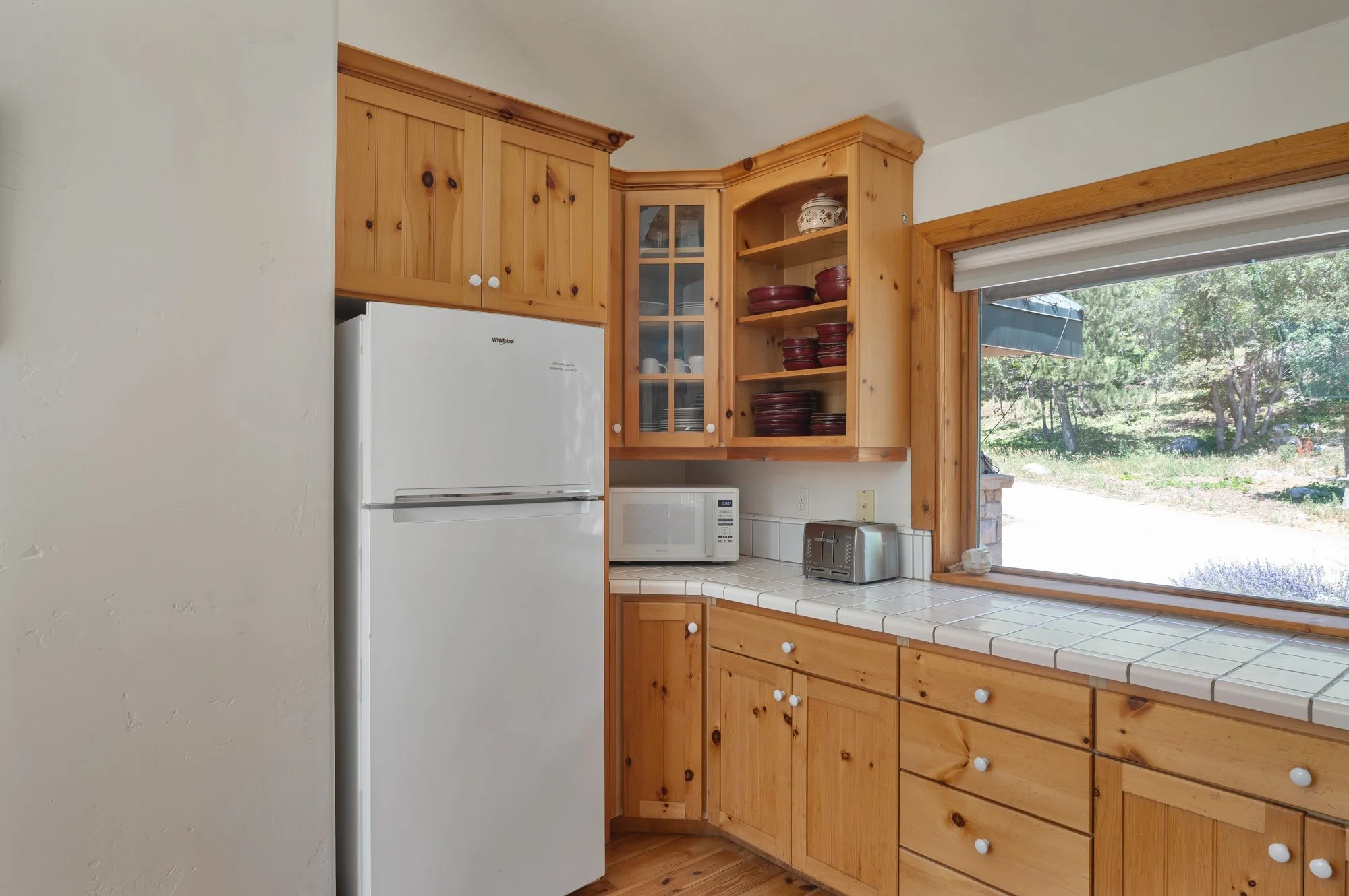 Kitchen with wooden cabinets, white refrigerator, microwave, toaster, and a large window overlooking greenery.