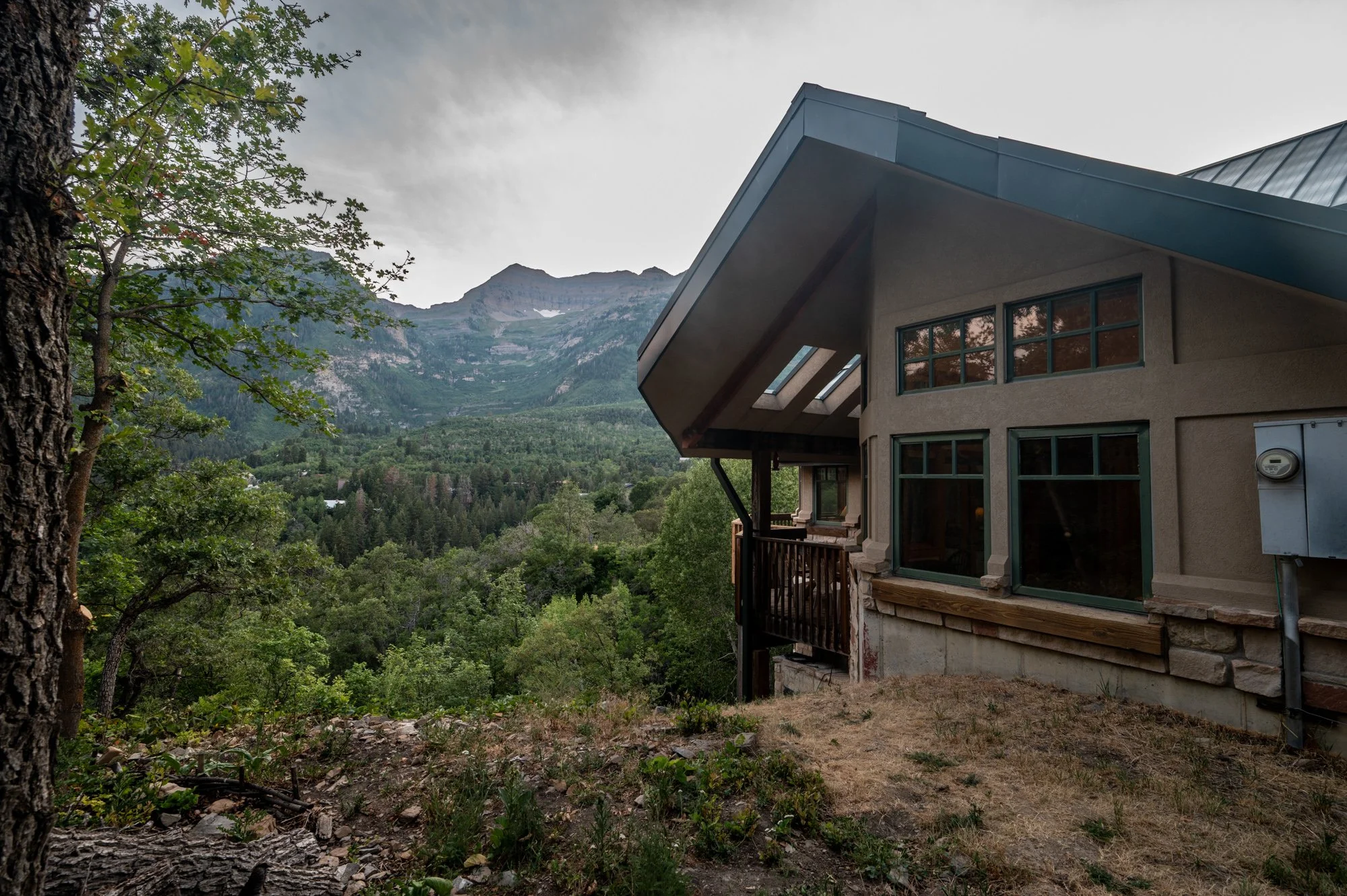 A house built on a hillside with large windows, a balcony, surrounded by green trees and mountains in the background under cloudy sky.