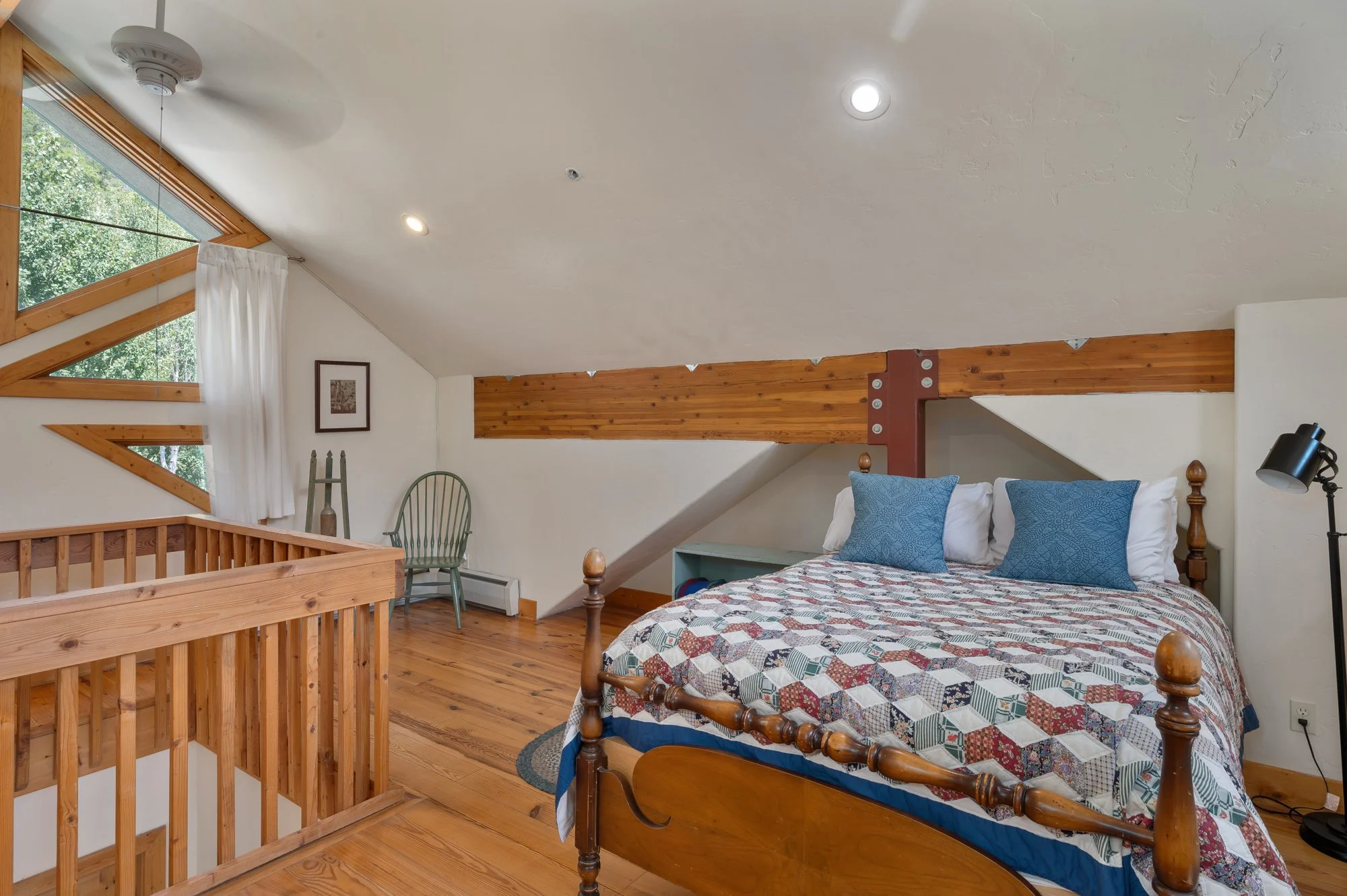 A cozy bedroom in an attic with a sloped ceiling, featuring a wooden bed with a colorful quilt and two blue pillows. Next to the bed is a small blue nightstand, and a black floor lamp is beside it. There is a large window with white curtains, a green