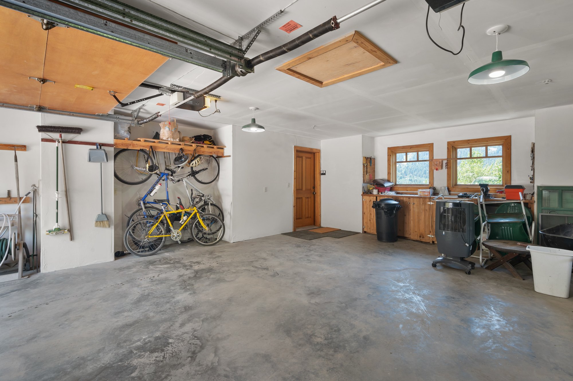 An organized garage with bicycles, tools, and storage shelves, featuring a concrete floor and two windows with a scenic view.