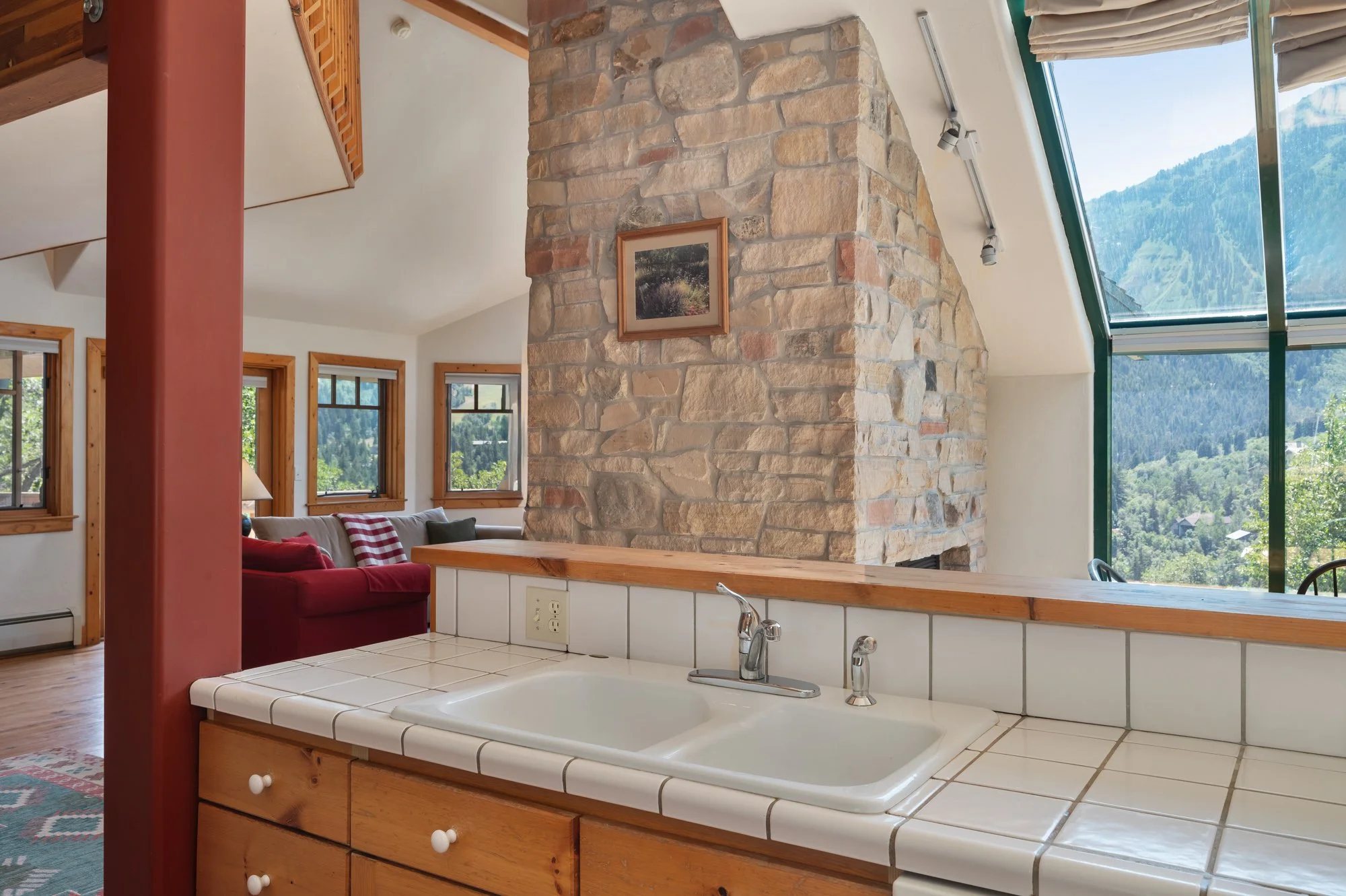 Kitchen with white tiled counter, double sink, and a large stone fireplace, overlooking a living area with windows showing a mountain view.