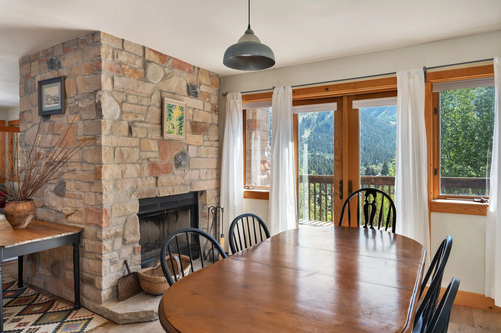 Interior of a cozy dining room with a large wooden table, black chairs, a stone fireplace, and a view of mountains outside through large windows with white curtains.