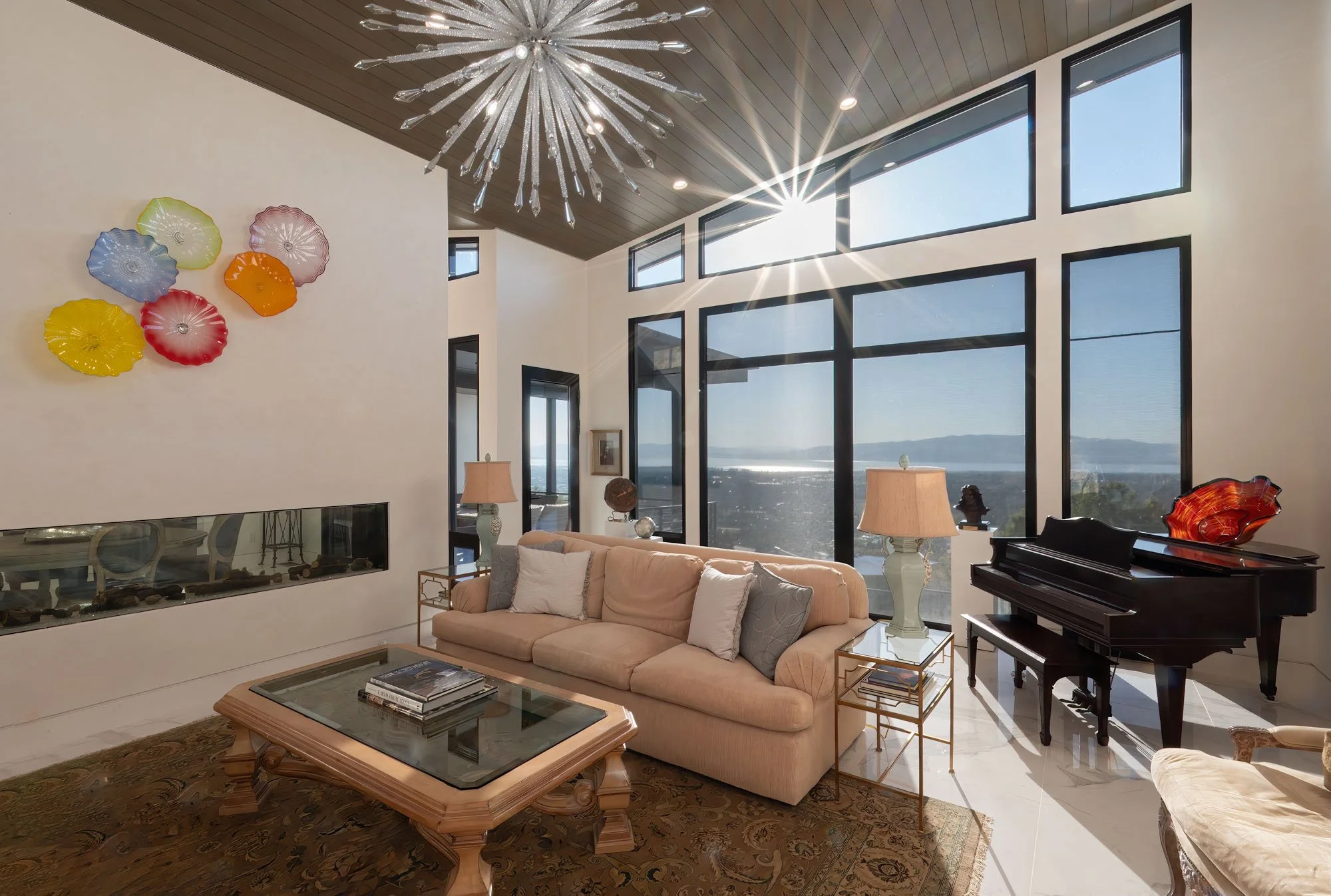 A spacious living room with large floor-to-ceiling windows overlooking a scenic view, modern art on the wall, a beige sofa with pillows, two side tables with lamps, a fireplace, a glass coffee table, a grand piano, and a colorful sculpture on top of the piano and wall.
