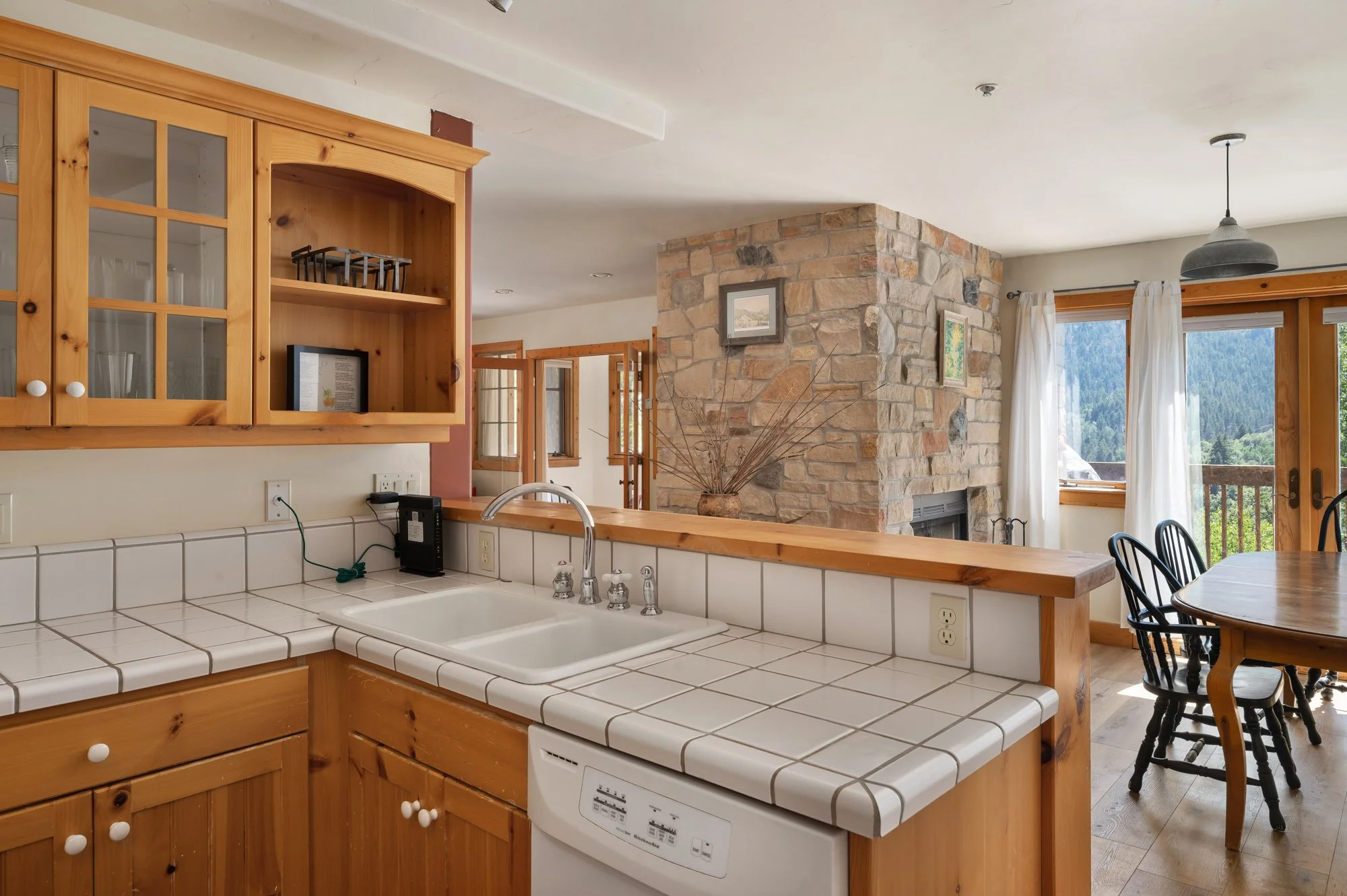 Kitchen with white tiled countertop, wooden cabinets, and a view into the dining and living room, which features a stone fireplace, windows with white curtains, and a balcony outside.