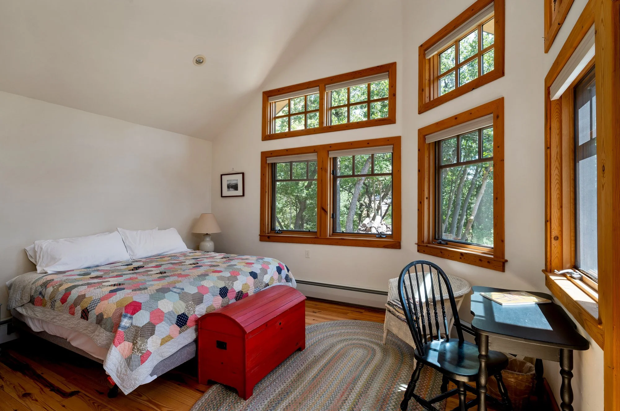 A cozy bedroom featuring a bed with a colorful patchwork quilt, a red wooden chest at the foot of the bed, a small white table lamp, a corner black desk with a black chair, and multiple large windows with wooden frames that look out onto trees.
