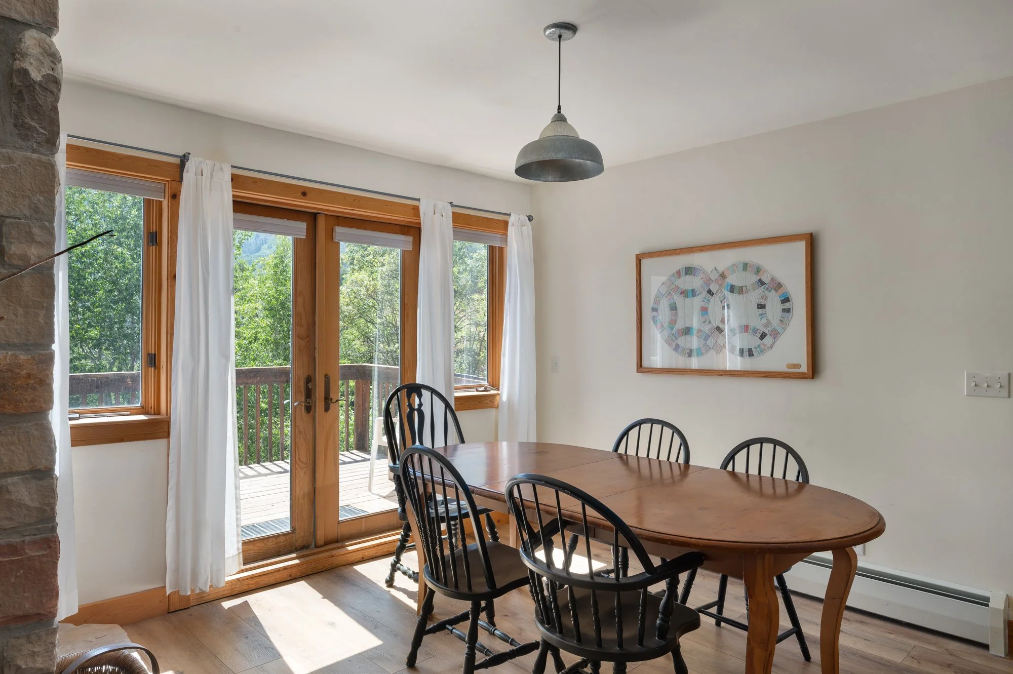 Dining room with a wooden table and four black chairs near large windows with white curtains, a framed artwork on the wall, and a sliding glass door leading to a balcony with trees outside.