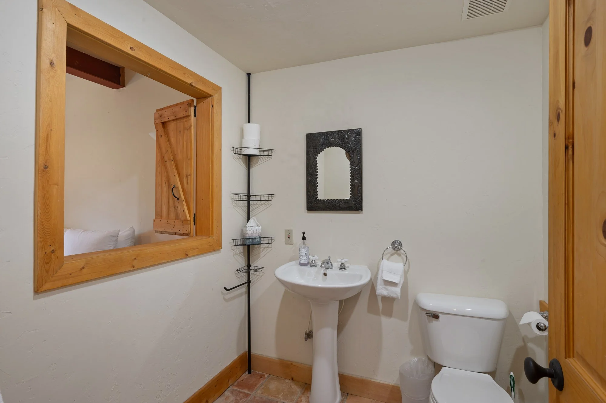 A small bathroom with a wooden-framed window looking into an adjacent room, a pedestal sink with soap, a towel ring with a towel, a toilet, a black decorative mirror, and wall-mounted metal shelves with toilet paper rolls and tissue box.