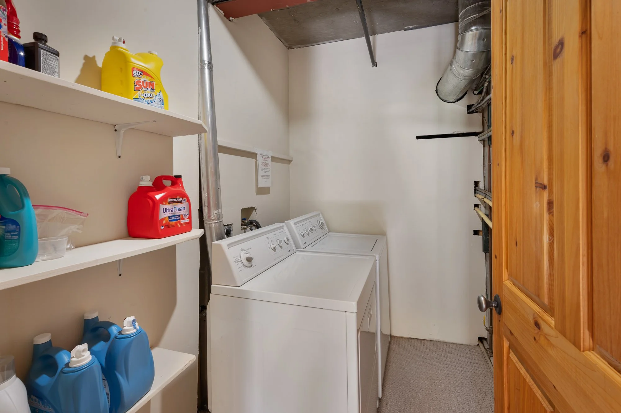 A laundry room with white washing machine and dryer, wooden door, and white shelves stocked with cleaning supplies including detergent and bleach.