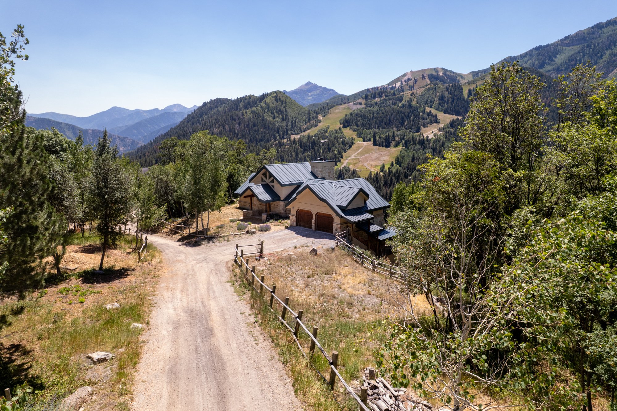 A house with a dark gray roof in a mountainous area surrounded by trees, with a dirt driveway leading up to it.