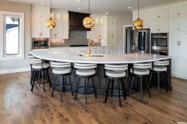 Modern kitchen with white cabinets, black appliances, central island with white countertop, gold pendant lights, and hardwood floors.