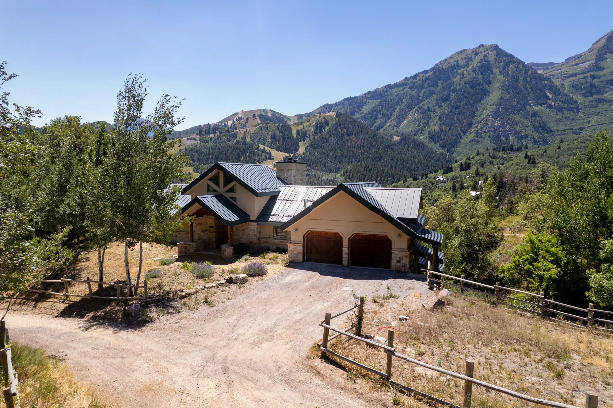 A house with a metal roof and stone accents, surrounded by trees and mountains in the background, on a sunny day.