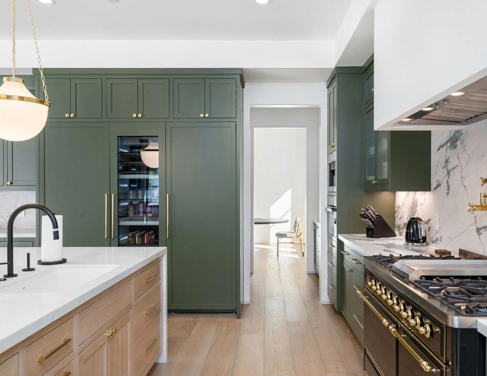 Modern kitchen with green cabinets, white marble countertops, a gold and black stove, black appliances, and a view into a dining area with a table and chairs.