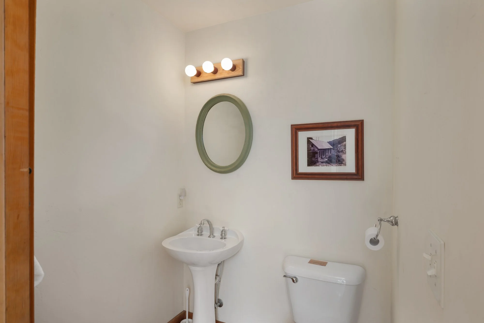 A small bathroom with a pedestal sink, an oval mirror, and a framed picture of an outdoor scene on the wall. There is a wooden light fixture with three bulbs above the mirror, and a toilet paper holder with a roll next to the toilet. The walls are pa