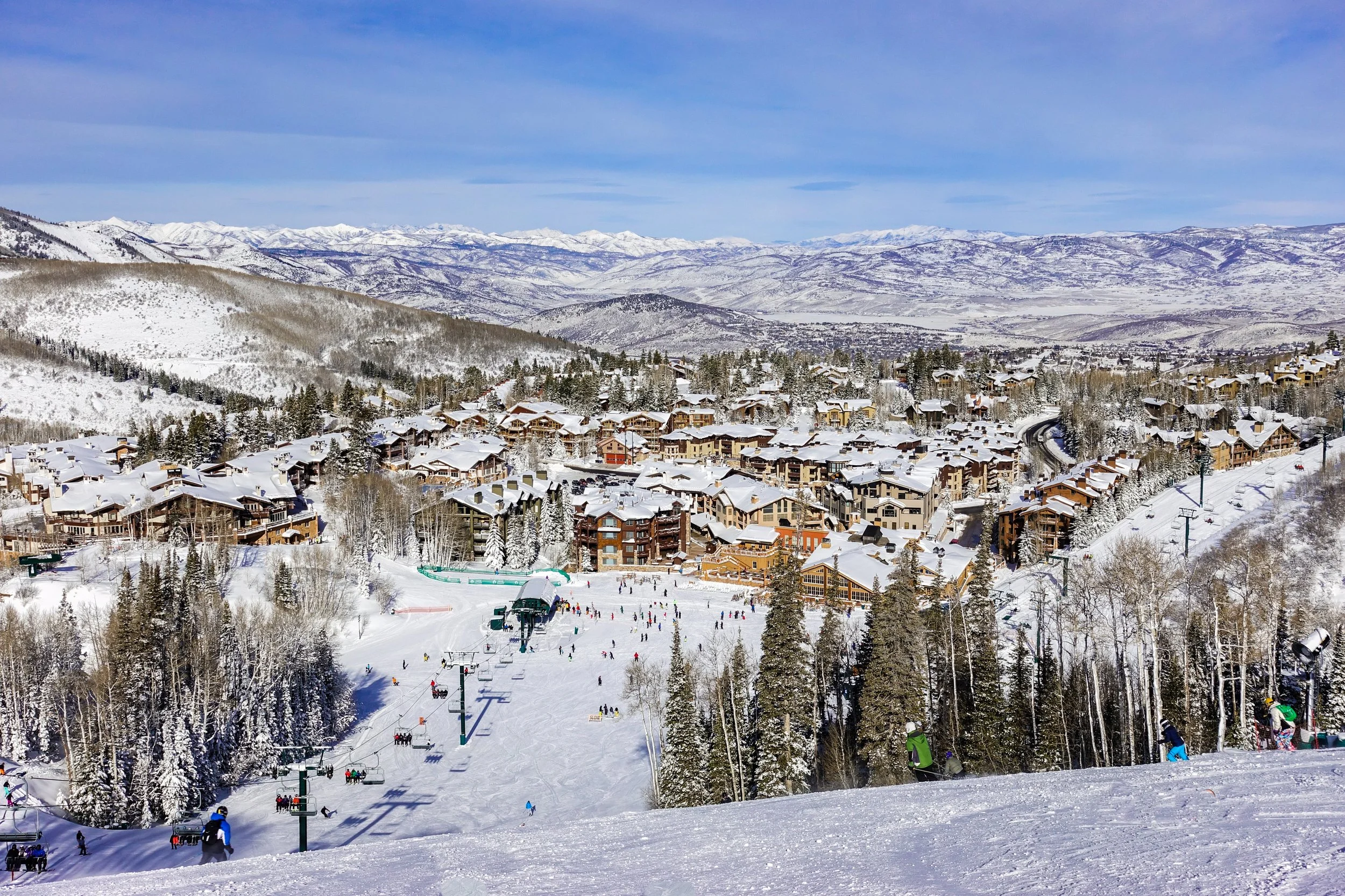 Snow-covered ski resort town with ski slopes, skiers, chair lifts, and mountain views in the background.