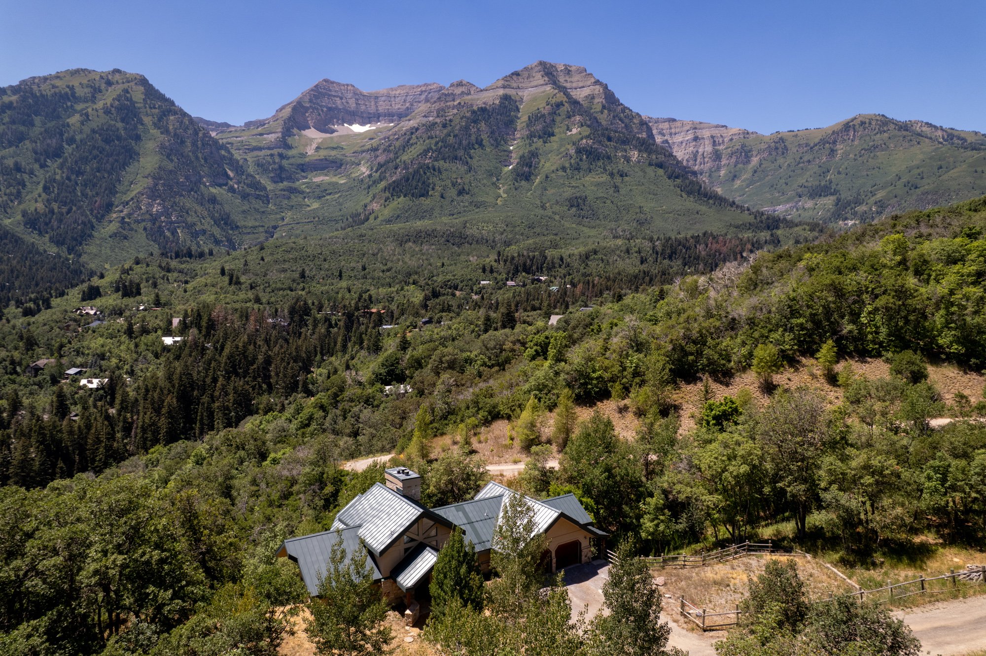 A house located in a lush, green, forested area with mountains in the background under a clear blue sky.