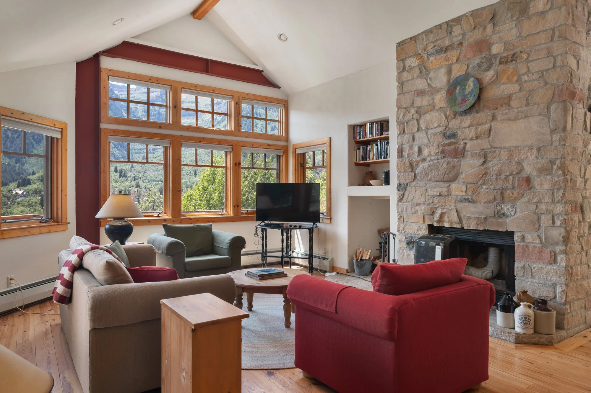 Living room with beige, green, and red armchairs arranged around a wooden coffee table, a stone fireplace, and large windows showing a mountain view.