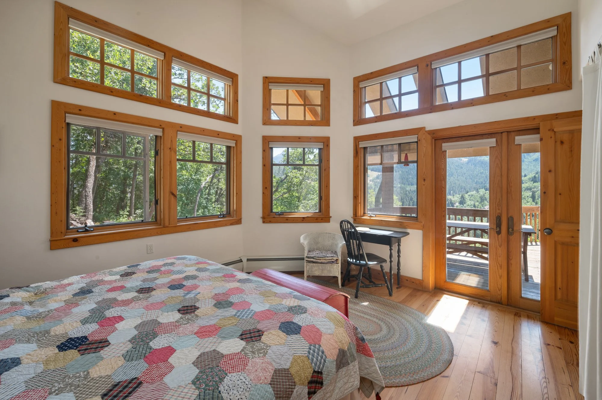 Sunlit bedroom with multiple large windows and a glass door leading to a balcony, a bed with a patchwork quilt, a small desk with a chair, a wicker chair, and a colorful oval rug, overlooking a mountain landscape.