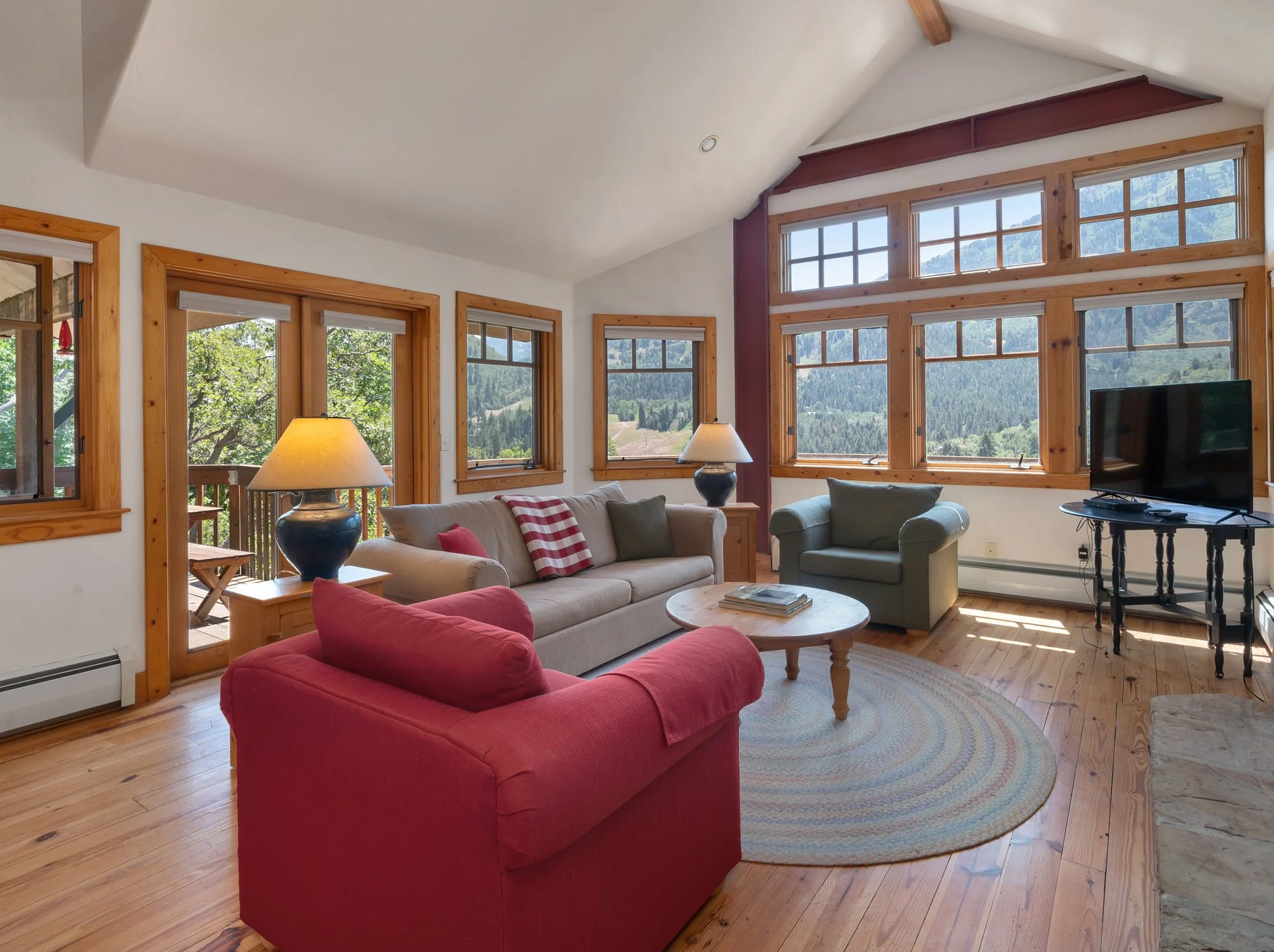 A living room with large wooden-framed windows showcasing a mountain view. It features a beige sofa with cushions, a red armchair, a round coffee table with books, two lamps on side tables, a TV on a black stand, and wood flooring with an oval rug.