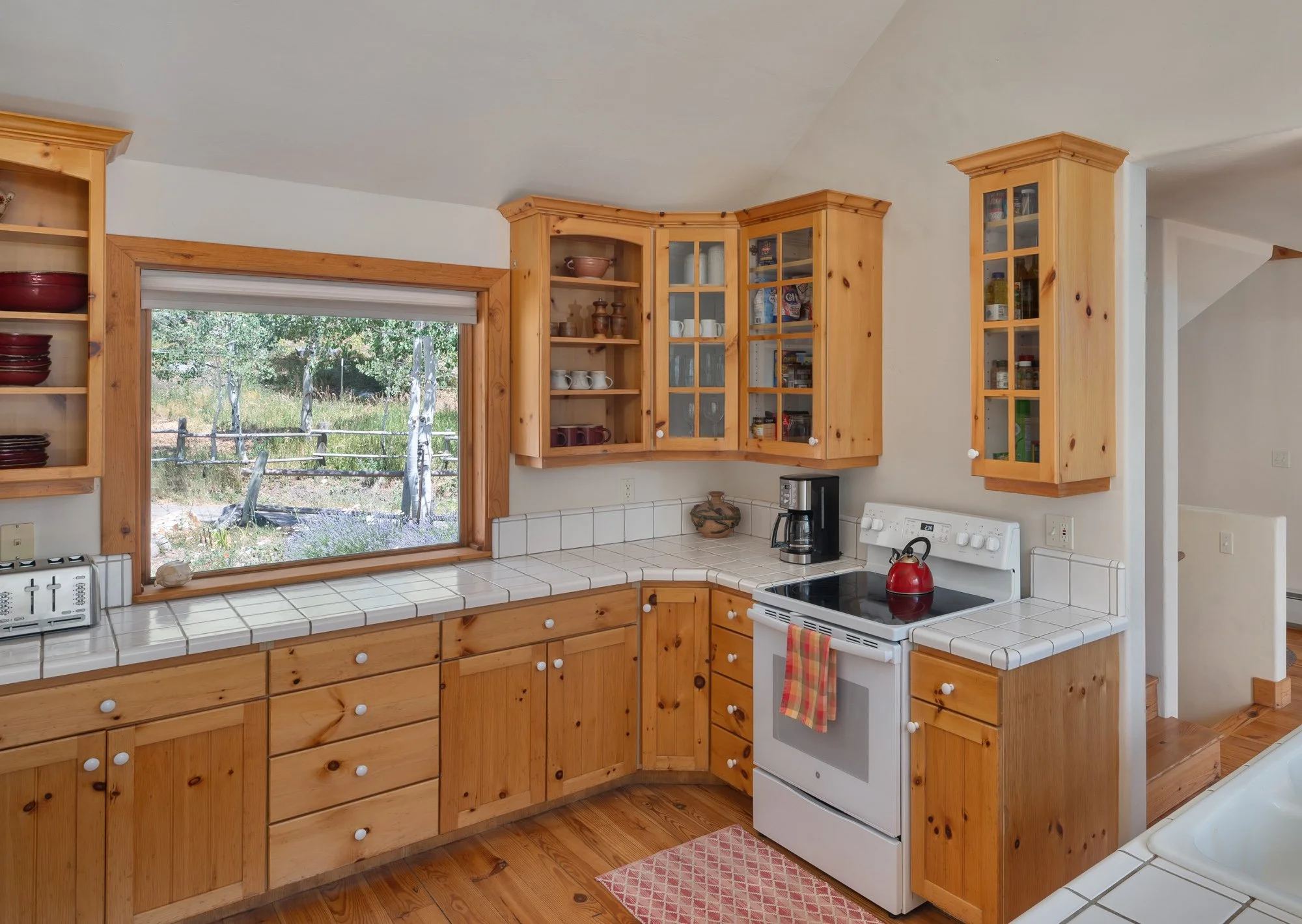 Wooden kitchen cabinets with white tiled countertops, a window showing an outdoor scene, a white stove with a red teapot, a coffee maker, and a small pink rug on a wooden floor.