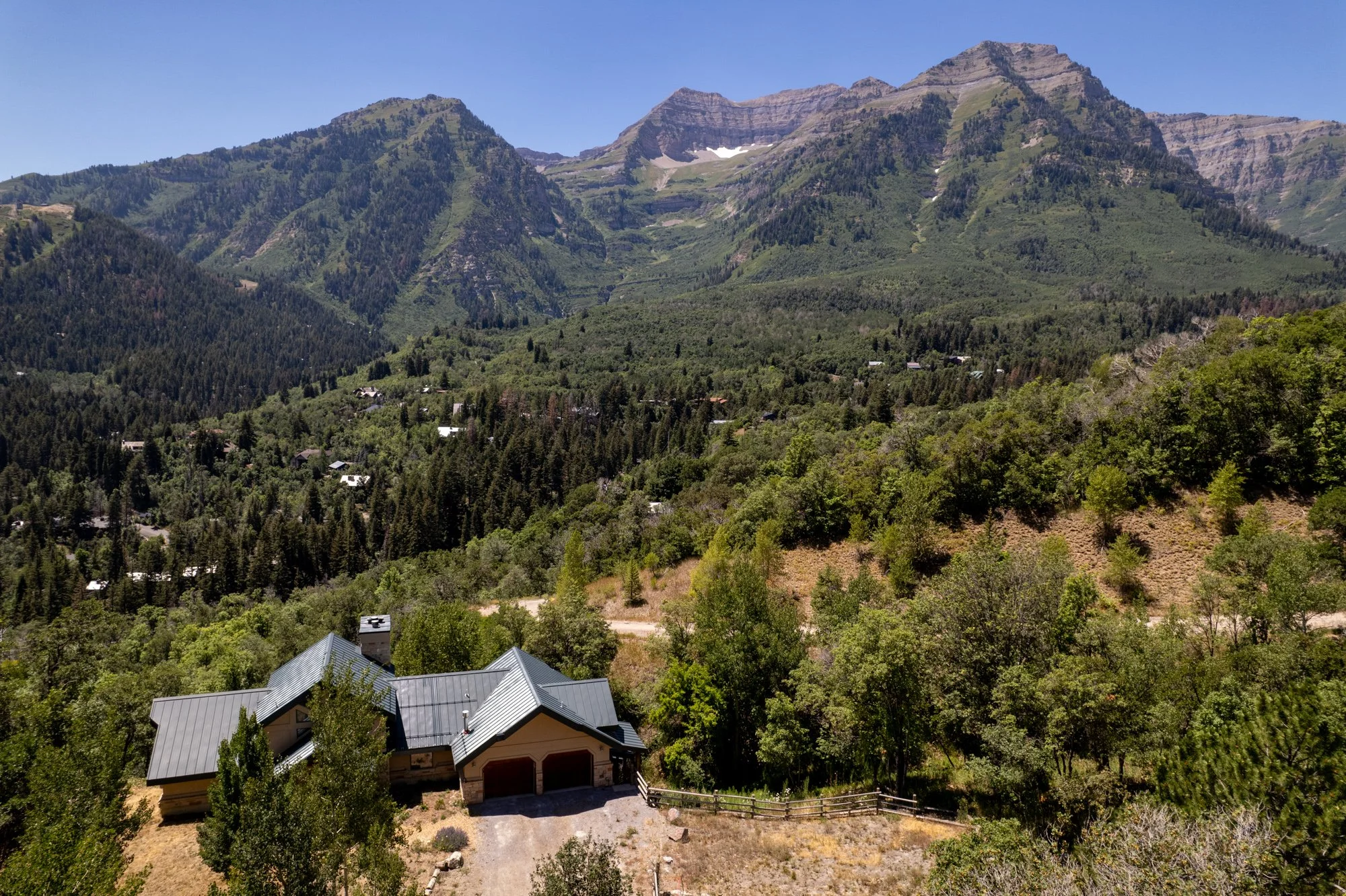 A house with a metal roof surrounded by trees, set against a mountain landscape with peaks, forest, and a clear blue sky.