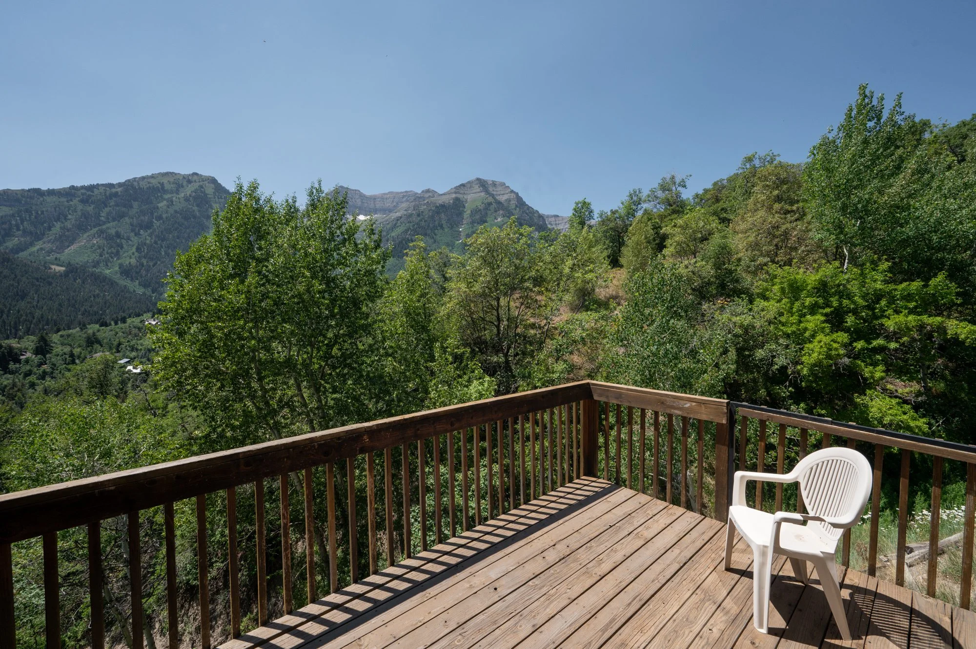 Wooden deck overlooking green trees and mountains on a clear, sunny day with a white plastic chair.
