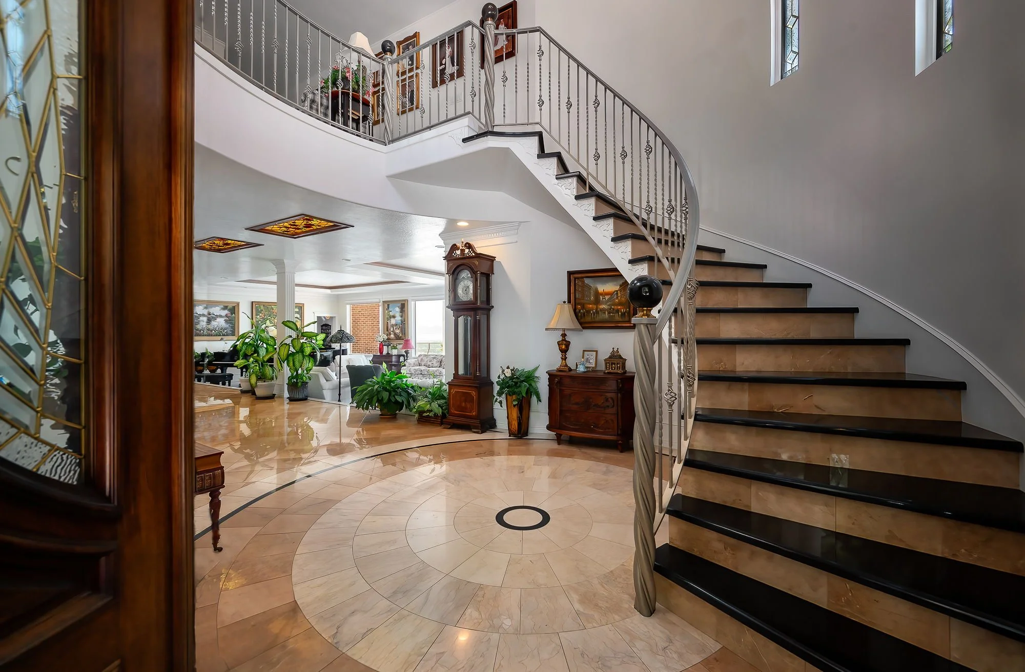 Interior of a spacious home featuring a curved staircase with beige steps and a metal railing, a hardwood floor with a circular design, and a foyer with antique furniture and plants. The background shows a living area with sofas, large windows, and decorative ceiling panels.