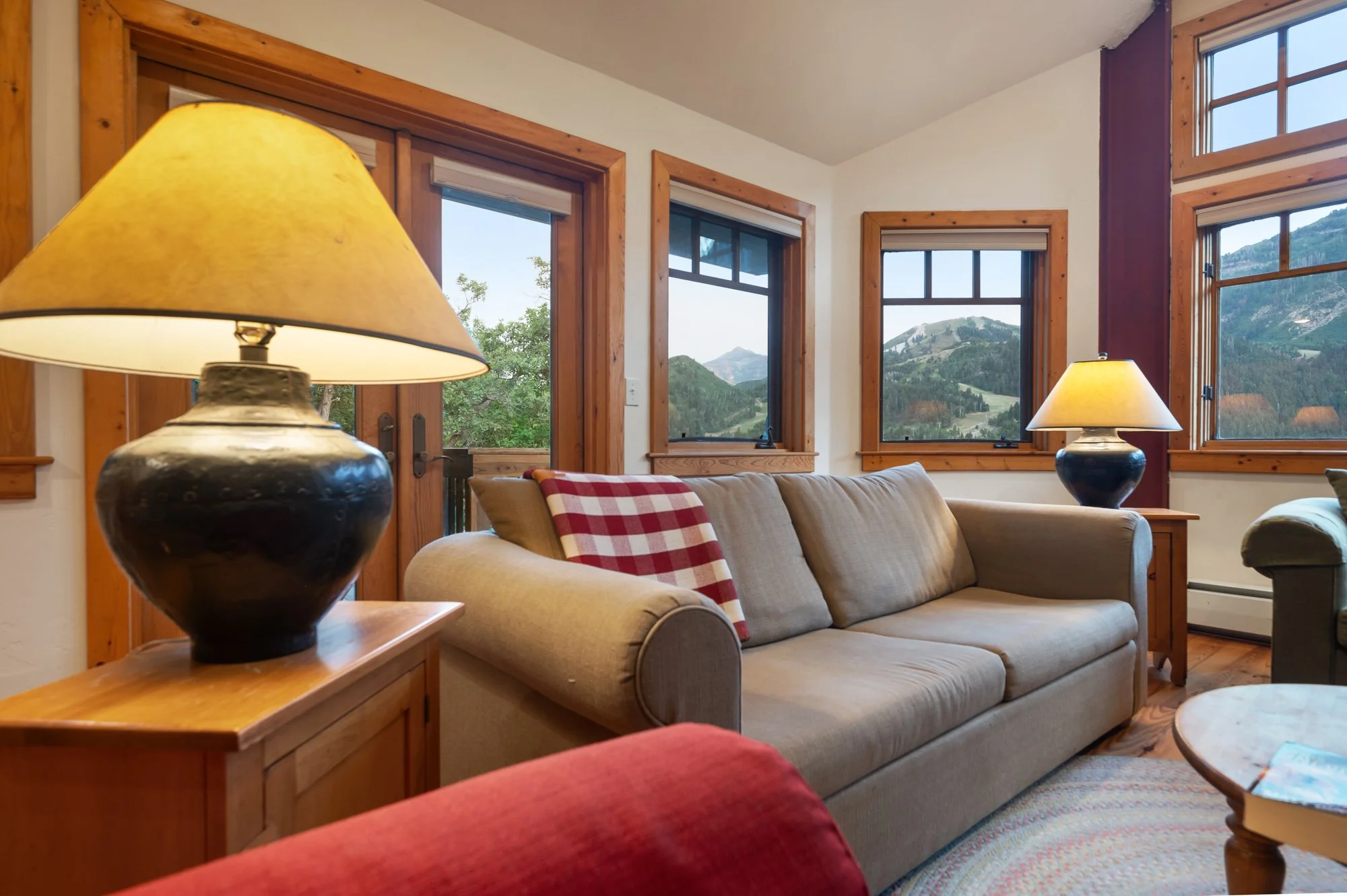 Living room with beige sofa, wooden window frames, and mountain view outside. Two lamps with yellow shades on side tables, and a red checkered blanket on the sofa.