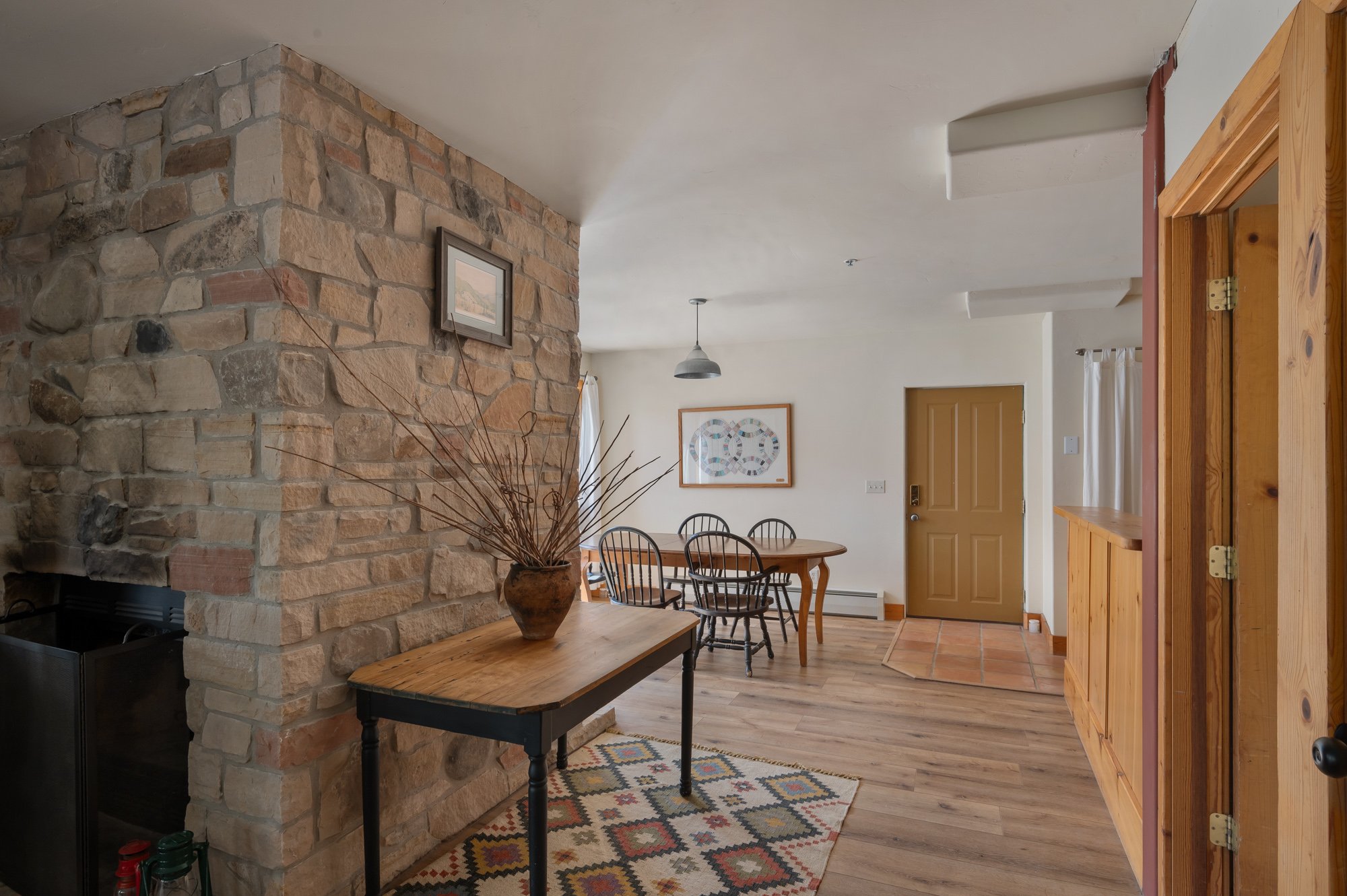 Interior of a home with a stone fireplace, wooden furniture, a dining table with chairs, hardwood floors, and a front door with a small entryway