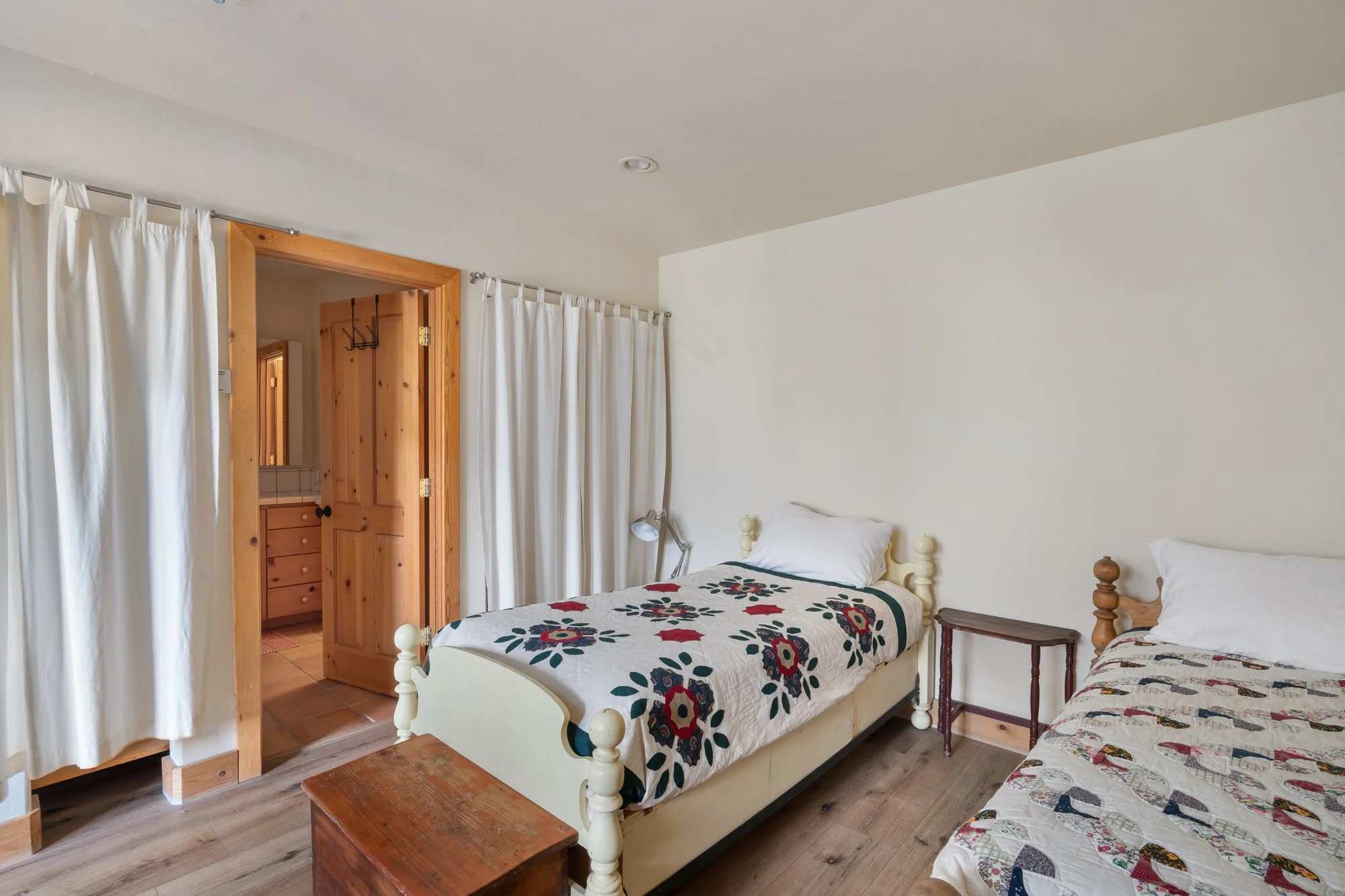 Hotel room with two beds, a small wooden side table, and a wooden doorway leading to a bathroom with a vanity and hooks.