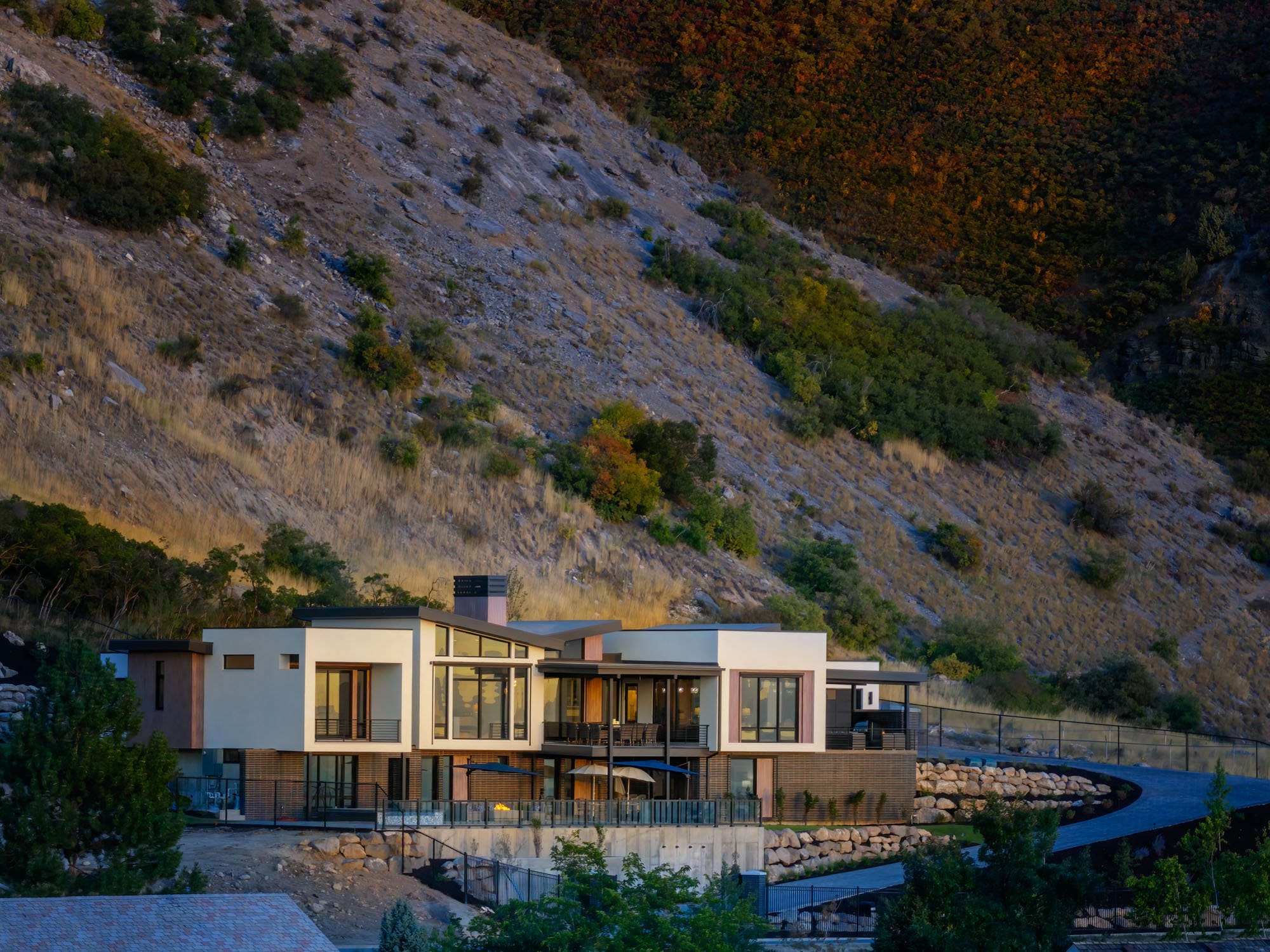 Modern house with large windows and multiple levels at the base of a rocky, wooded hillside.
