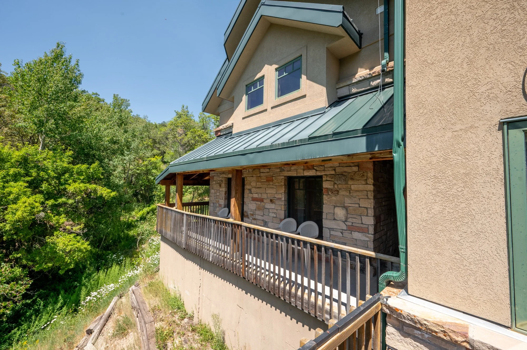 Balcony of a house with a stone and stucco exterior, green metal roof, and wooden railing. Two chairs are on the balcony, overlooking lush green trees and a blue sky.