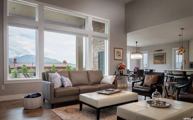 Bright living room with large windows, gray sofa, black armchair, white ottoman, and view of mountains and houses outside.