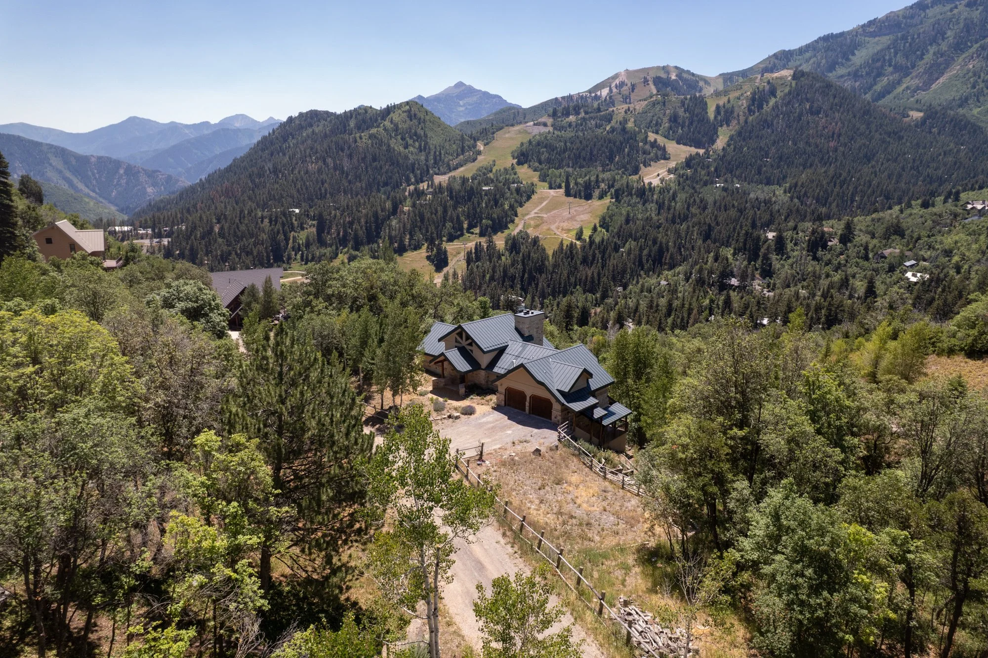 A house in a forested mountainous area with a driveway, surrounded by trees and hills, under a clear sky.