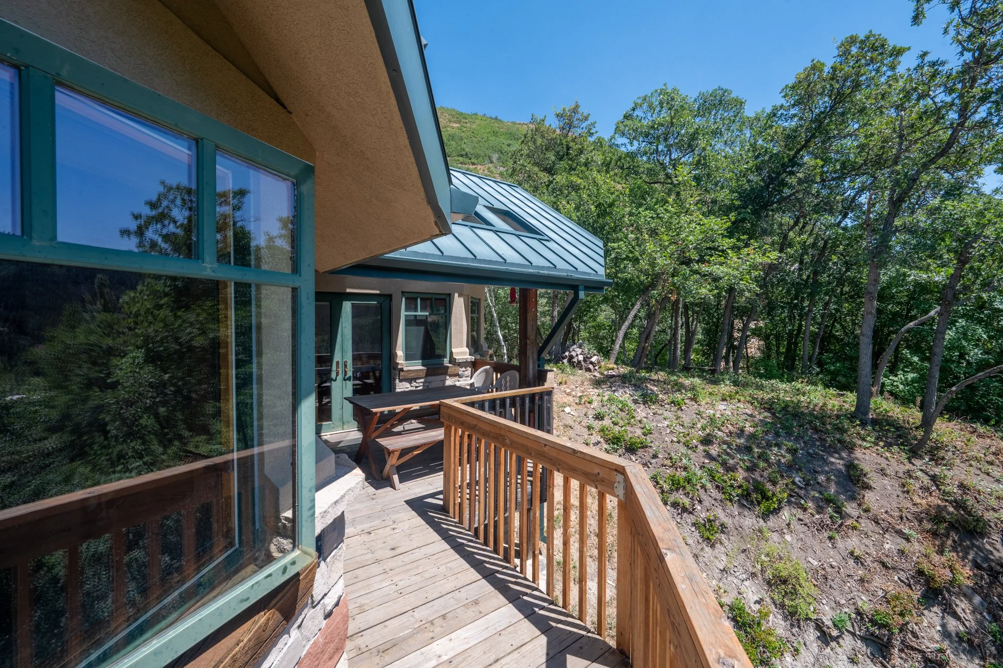 Wooden balcony with railing attached to a house with large windows, overlooking trees and a hillside under a clear blue sky.