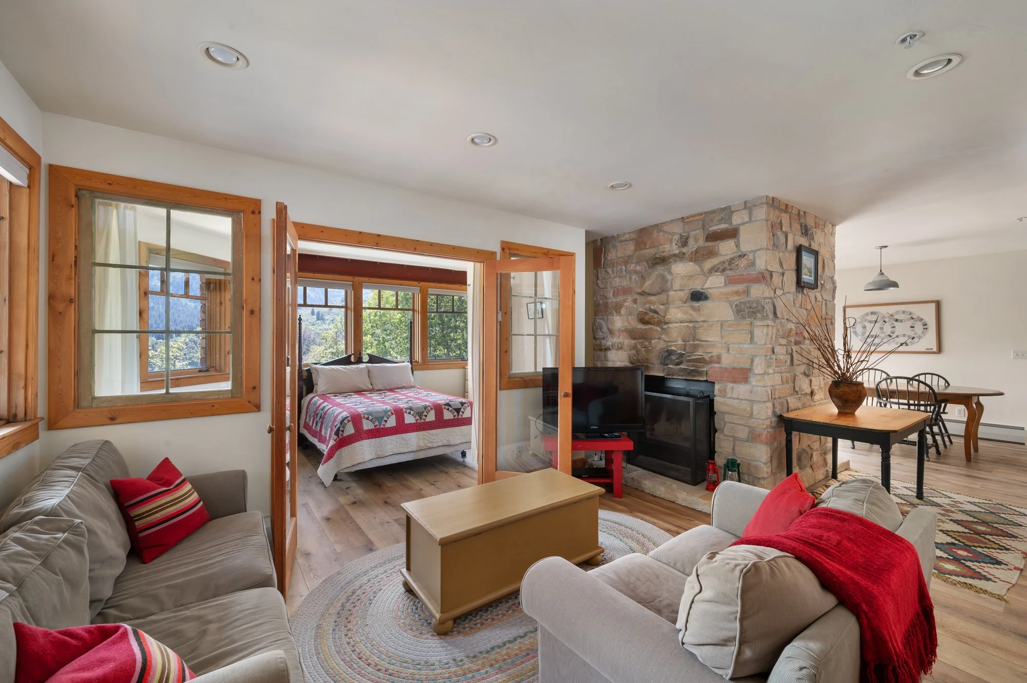 Living room with beige sofas and red cushions, a wooden coffee table, a stone fireplace, and a view of an adjoining bedroom with a bed and windows showing a mountain landscape.