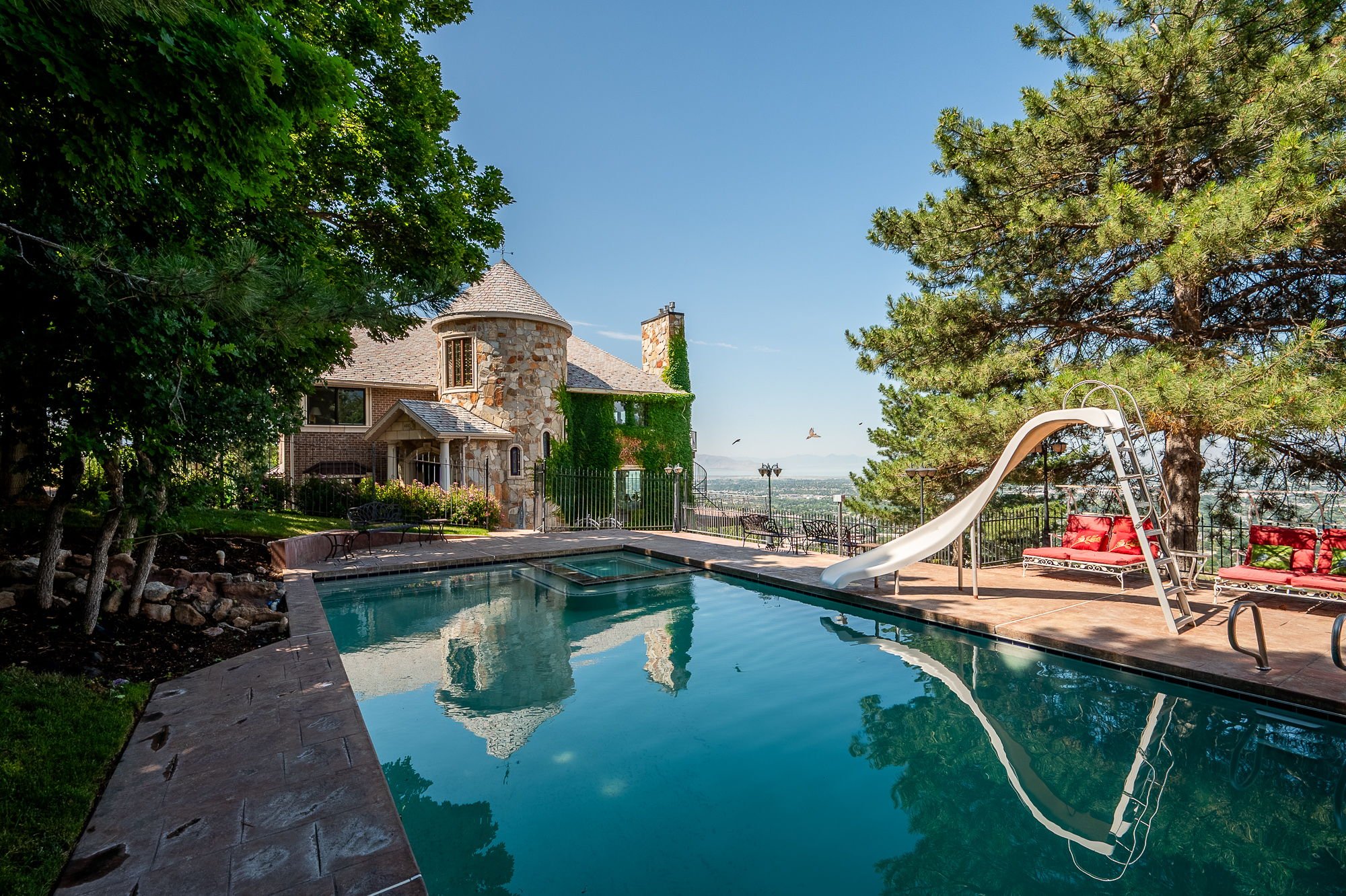 Residential backyard with swimming pool, small slide, sitting area with red cushions, large trees, a stone house, and a view of the city skyline in the background.