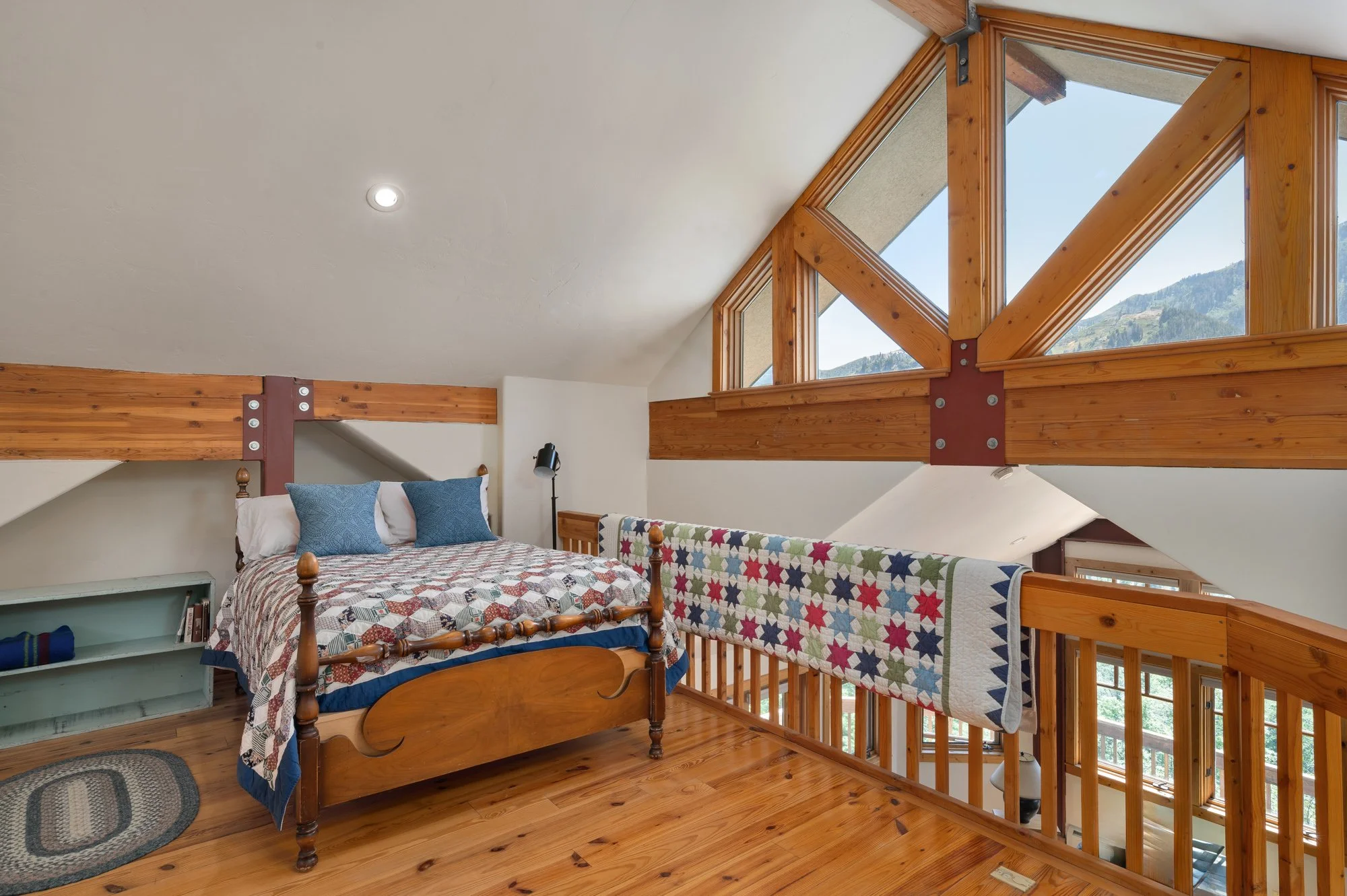 Interior of a cozy loft bedroom with a wooden bed, colorful quilt, two blue pillows, a black reading lamp, and a mountain view through large triangular windows.