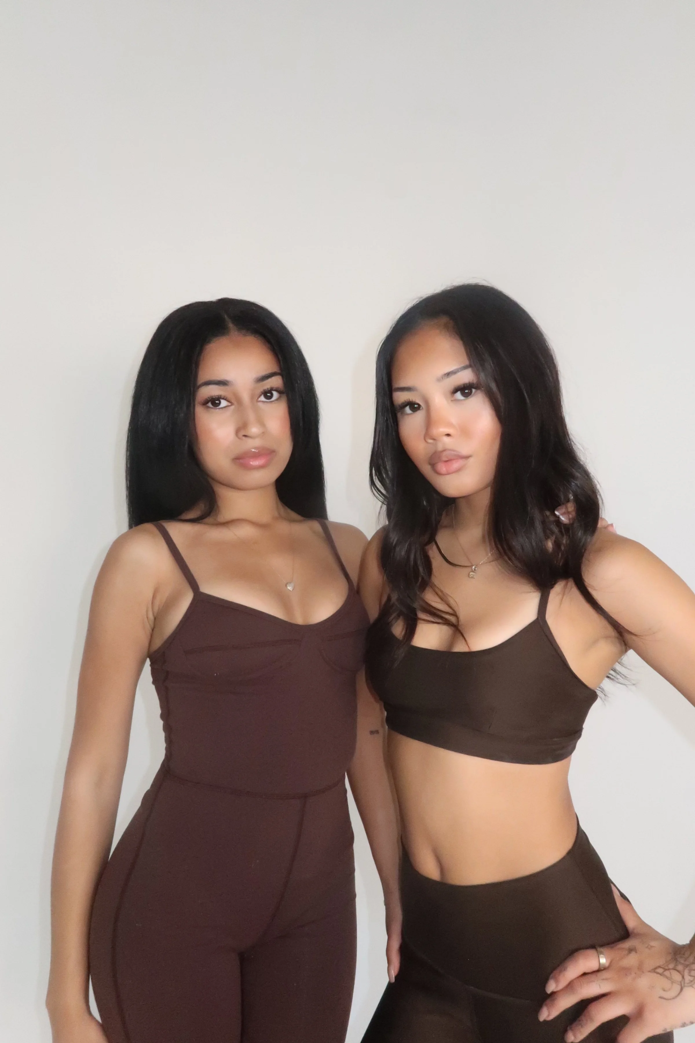Two young women standing together against a plain white wall, both wearing dark brown tops and pants, with natural makeup and shoulder-length hair.
