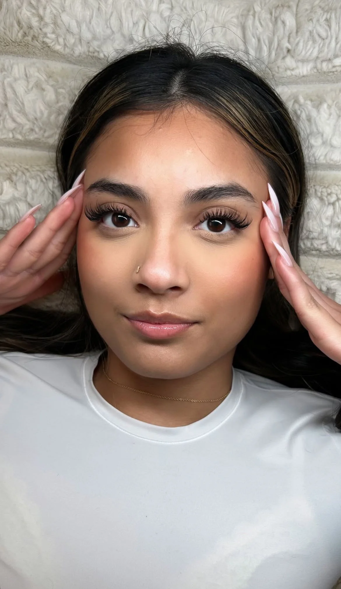 A young woman with long dark hair, wearing a white top and a delicate necklace, is touching her temples with both hands, looking directly at the camera, with a relaxed expression, and is standing in front of a light-colored textured wall.