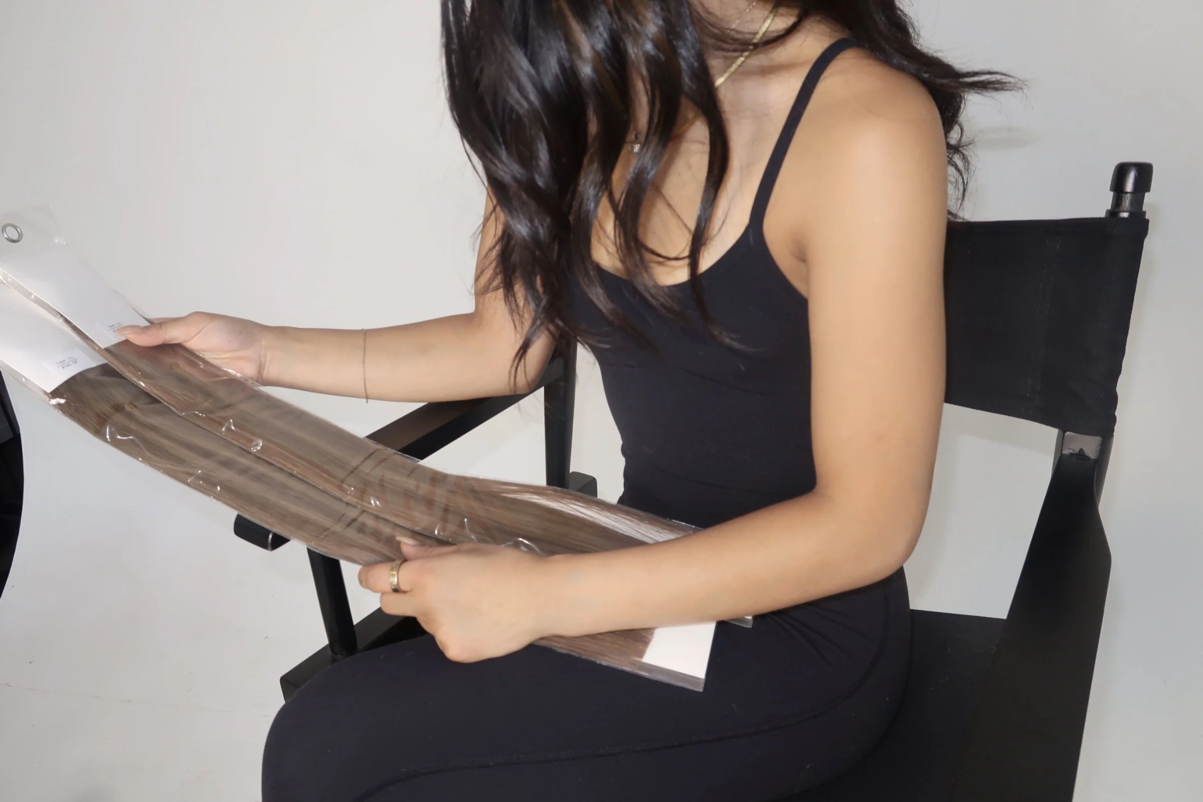 Woman in black top and black pants sitting on a black chair, holding a package of hair extensions.