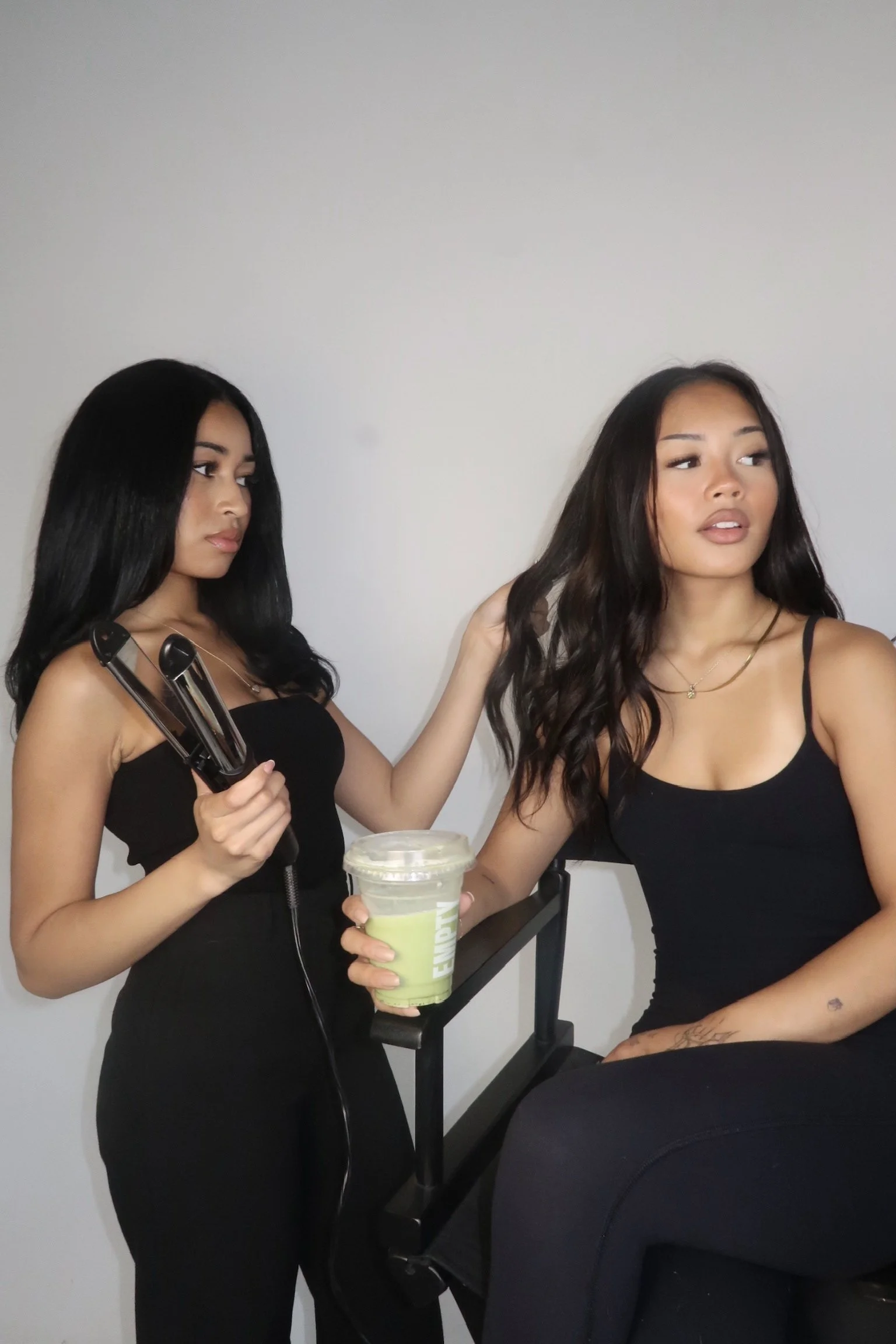 Two women getting their hair styled, one using a flat iron, with a neutral background.