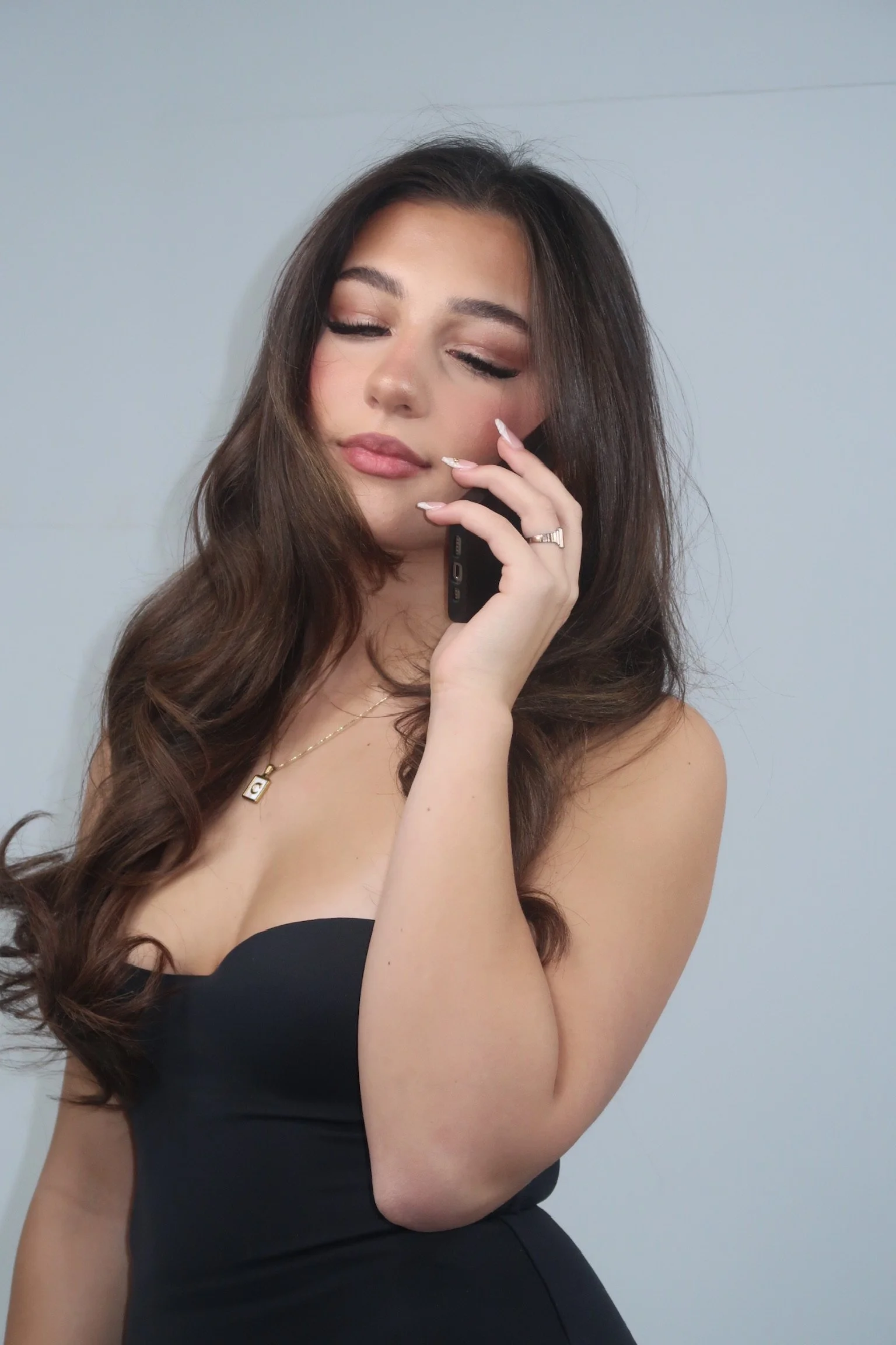 A young woman with long, wavy brown hair and makeup, wearing a black strapless dress, holding a phone to her ear, with her eyes closed and a relaxed expression.