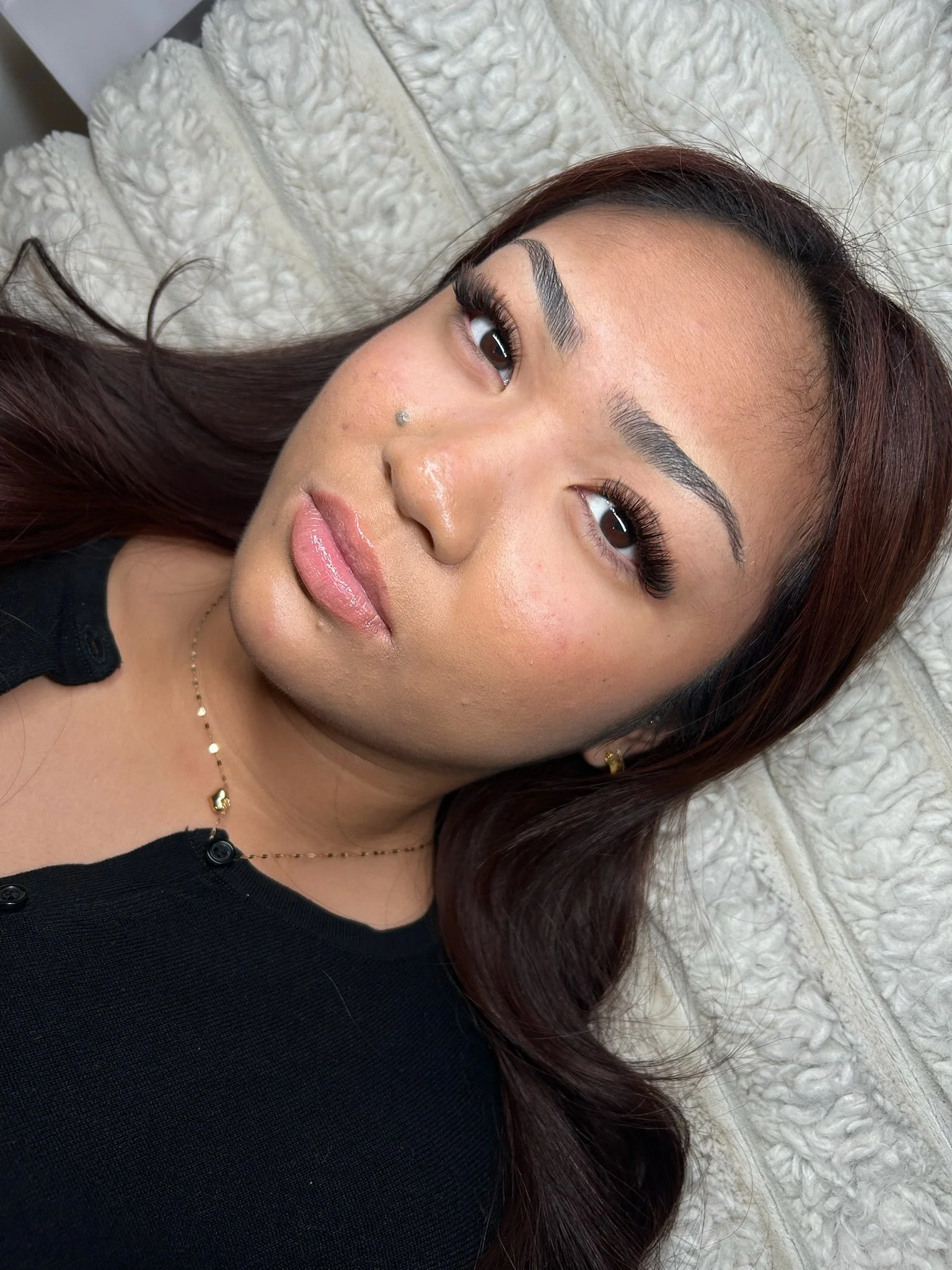 A woman with long brown hair, full eyebrows, makeup, and a nose piercing, lying on a textured white surface, looking at the camera.