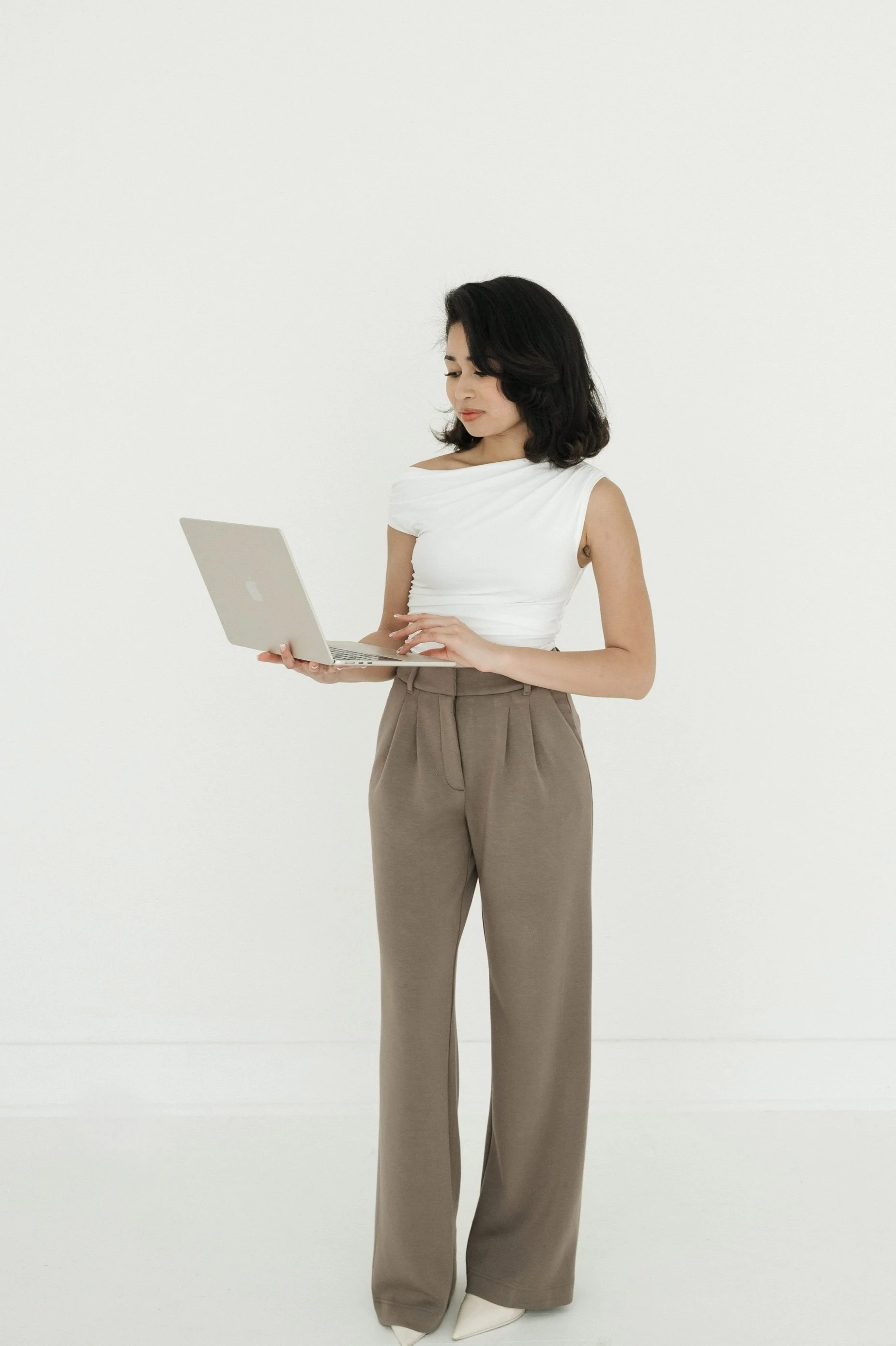 A woman with shoulder-length black hair wearing a white sleeveless top and tan high-waisted wide-leg pants, holding a silver laptop, standing against a plain white background.