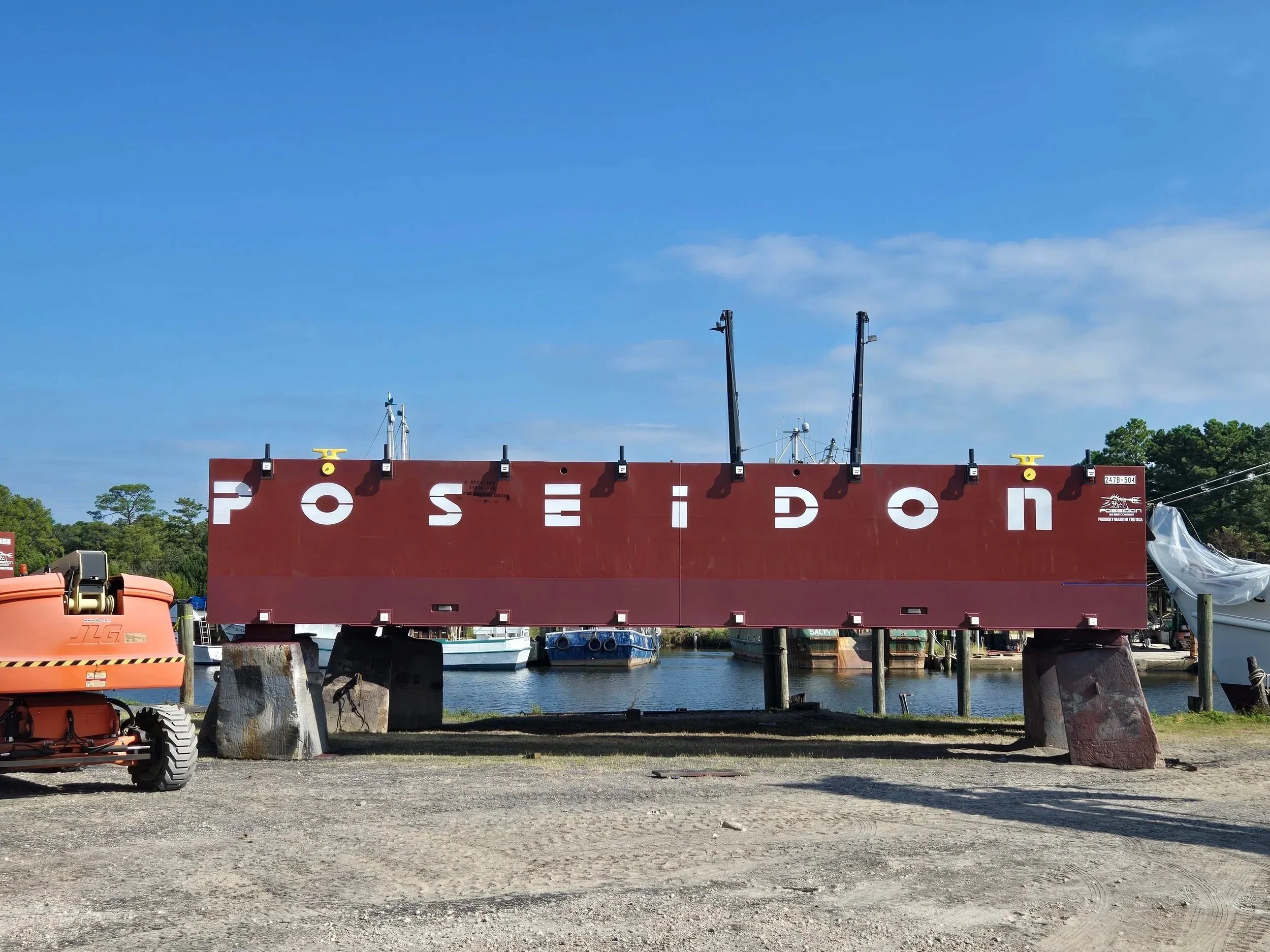 A large red sign on the ground with the word 'POSEIDON' painted in white letters, situated outdoors near a marina with boats and water in the background.