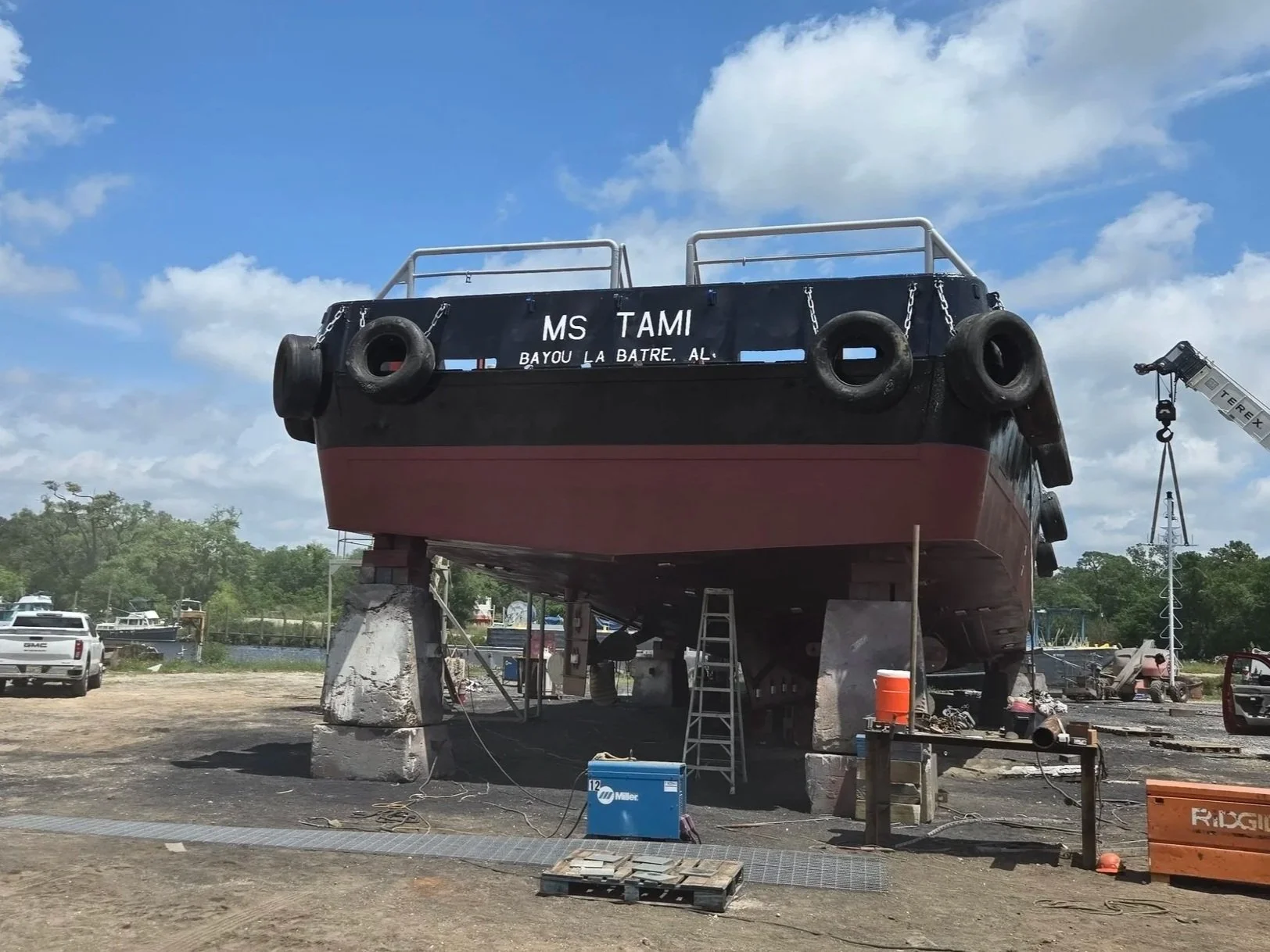 A large boat named MS TAMI is under construction on land, supported by concrete blocks and scaffolding, with tools and equipment around it, in a boatyard with a crane in the background.
