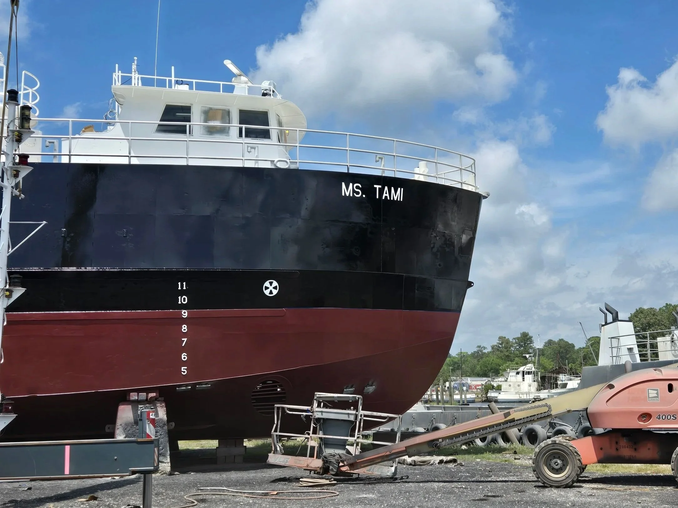 Large black and red ship named MS. Tami in a dry dock with a construction lift, tires, and equipment around it, under a partly cloudy sky.